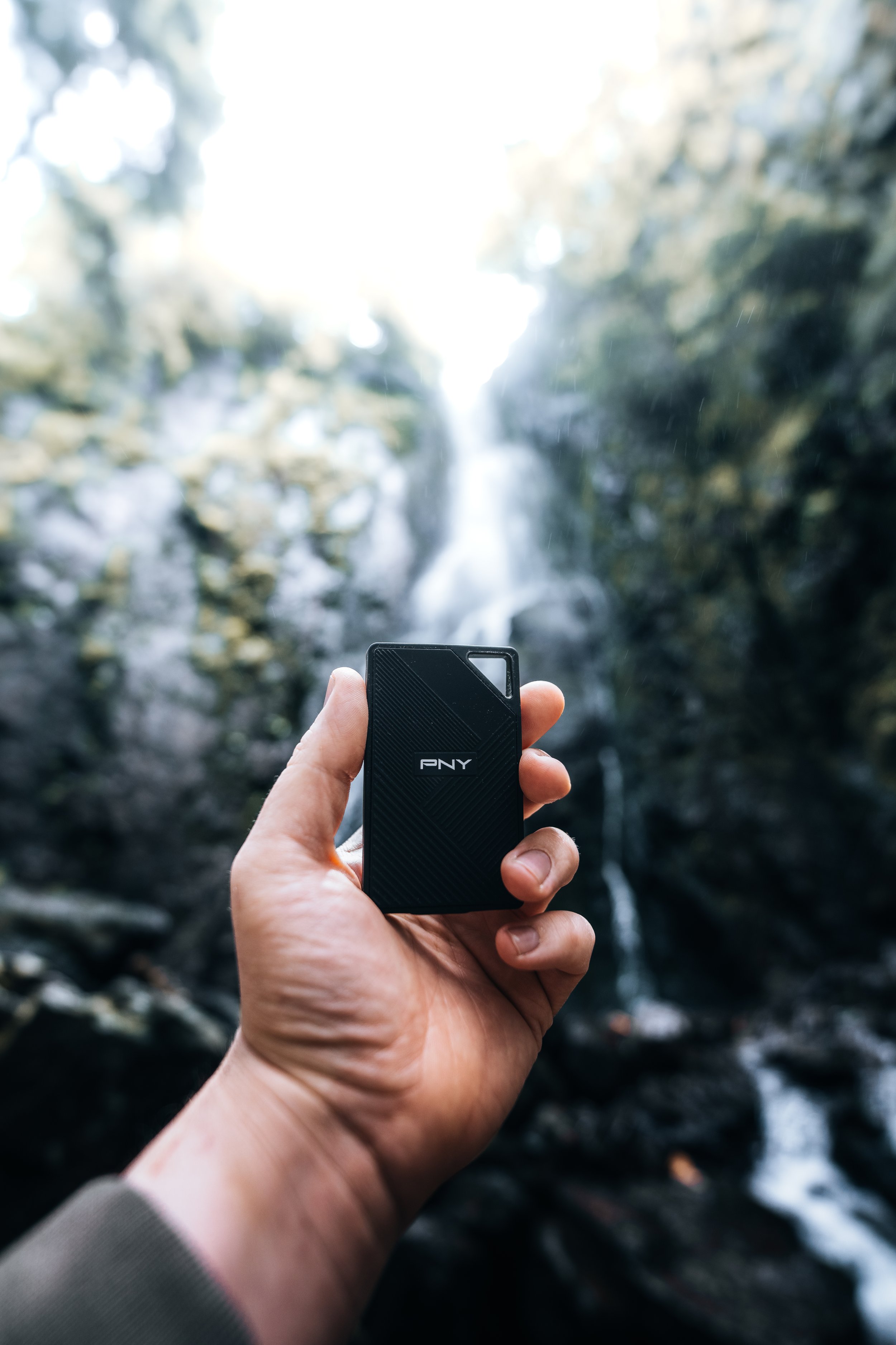 Hand hält einen kleinen schwarzen PNY externen Speicher vor einem Wasserfall in einer natürlichen, bewaldeten Umgebung.
