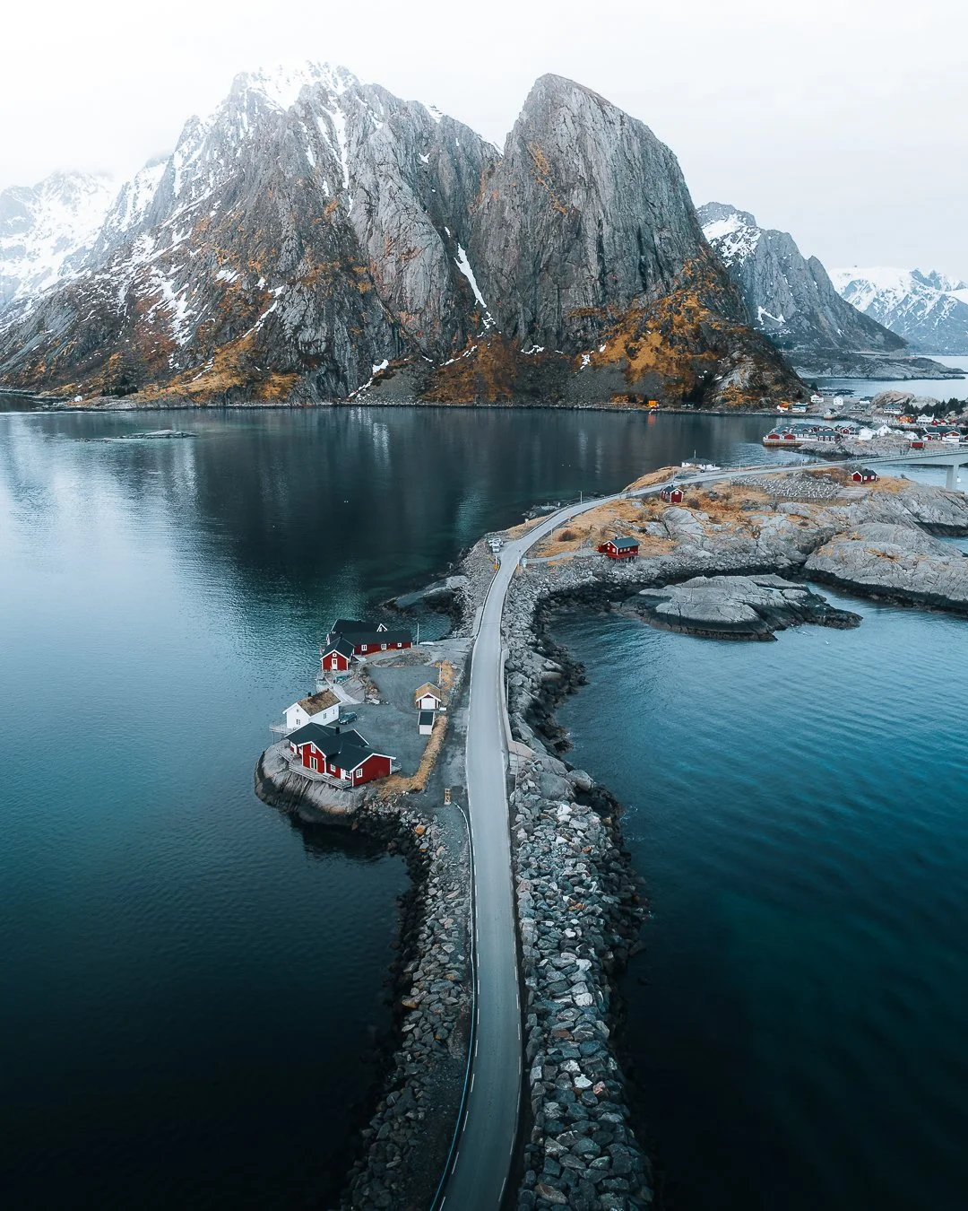 Blick auf eine schmale Straße, die eine Insel mit roten und weißen Häusern mit Blick auf einen ruhigen Fjord verbindet, umgeben von hohen, schneebedeckten Bergen in Norwegen.
