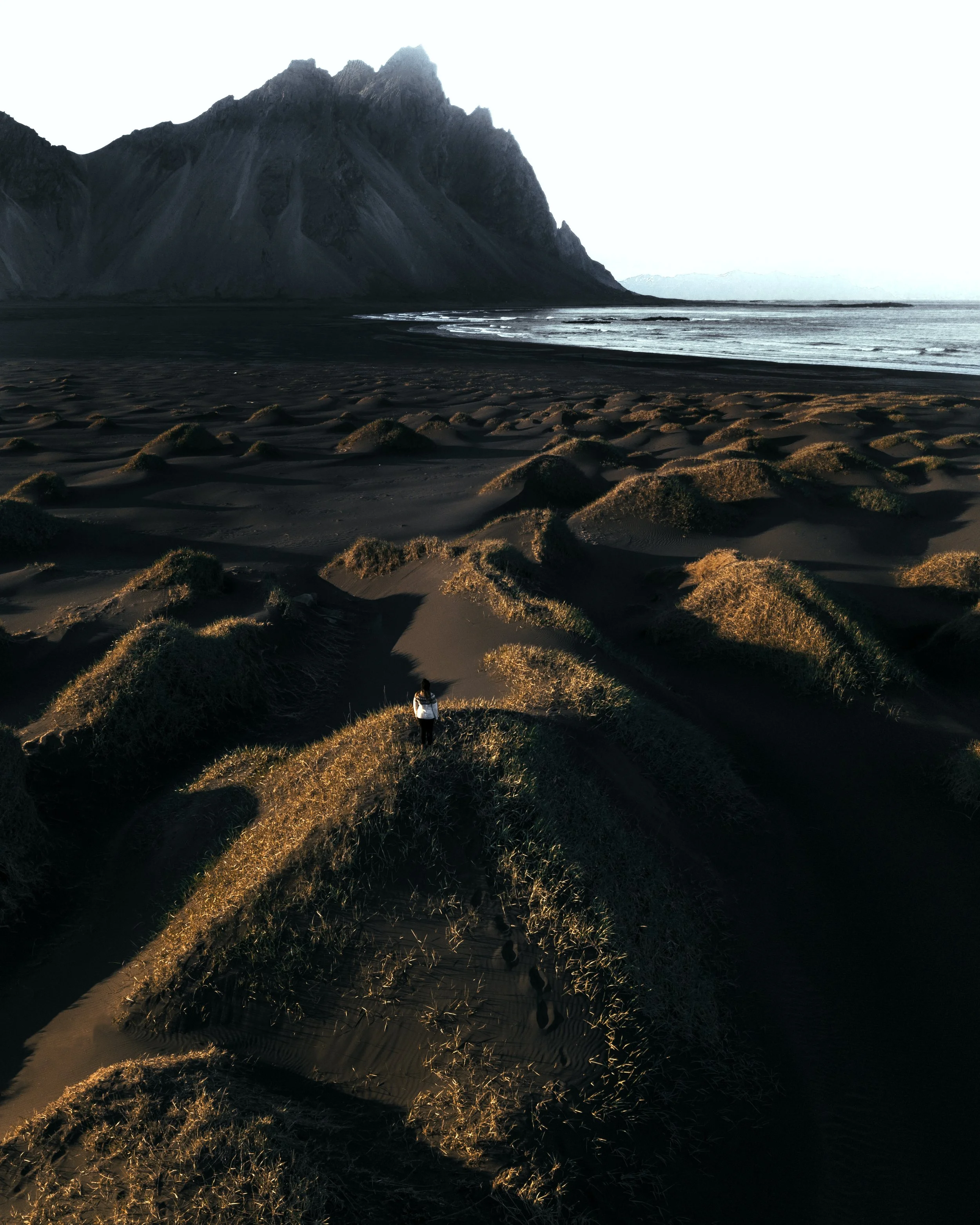 Eine einsame Person steht auf einer Düne in einer Wüstenlandschaft mit hohen Sandhügeln, im Hintergrund das Meer und ein Berg bei Sonnenuntergang.