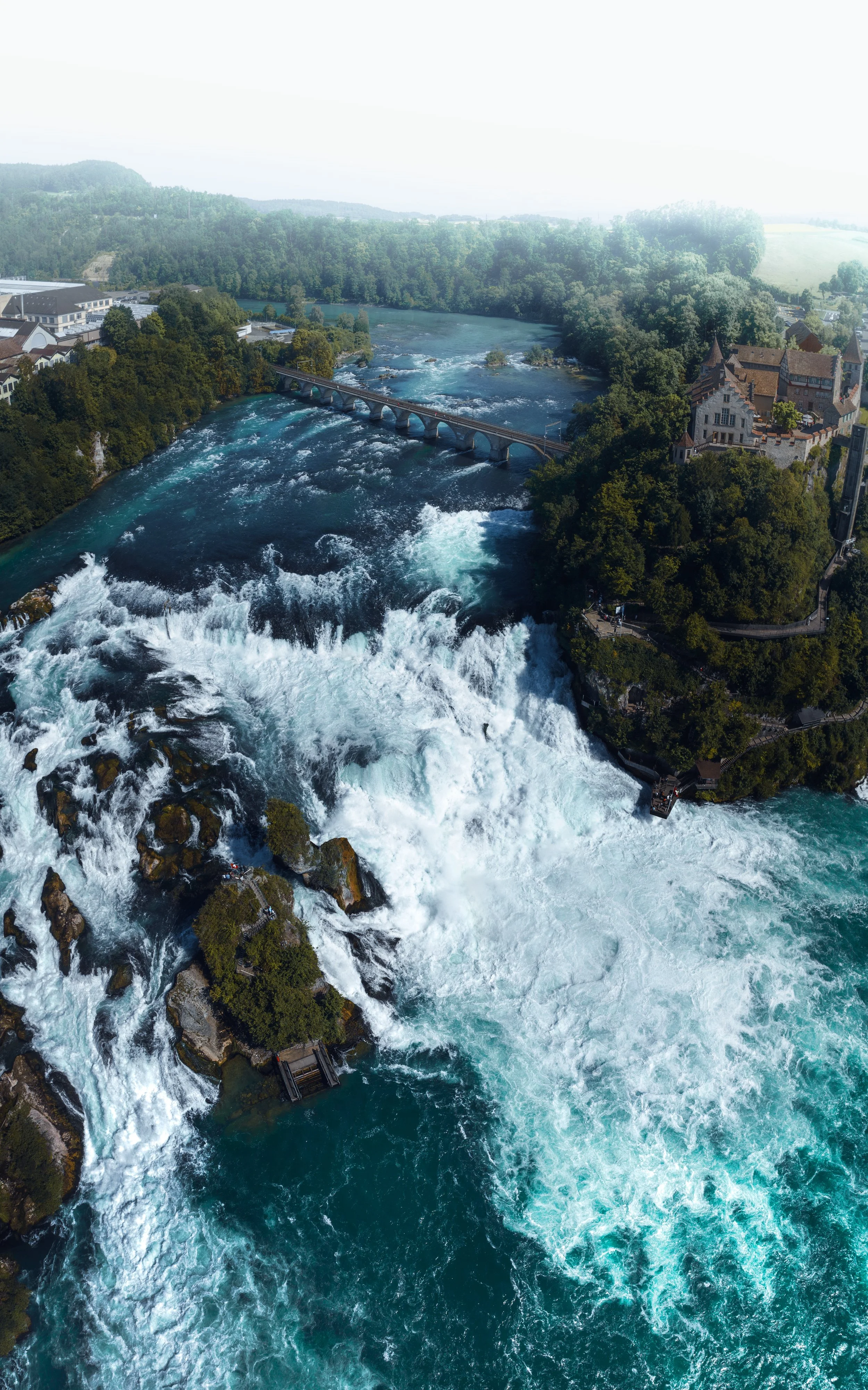 Luftaufnahme eines Flusses mit Wasserfall, umgeben von Bäumen und historischen Gebäuden, mit Brücke über den Fluss.