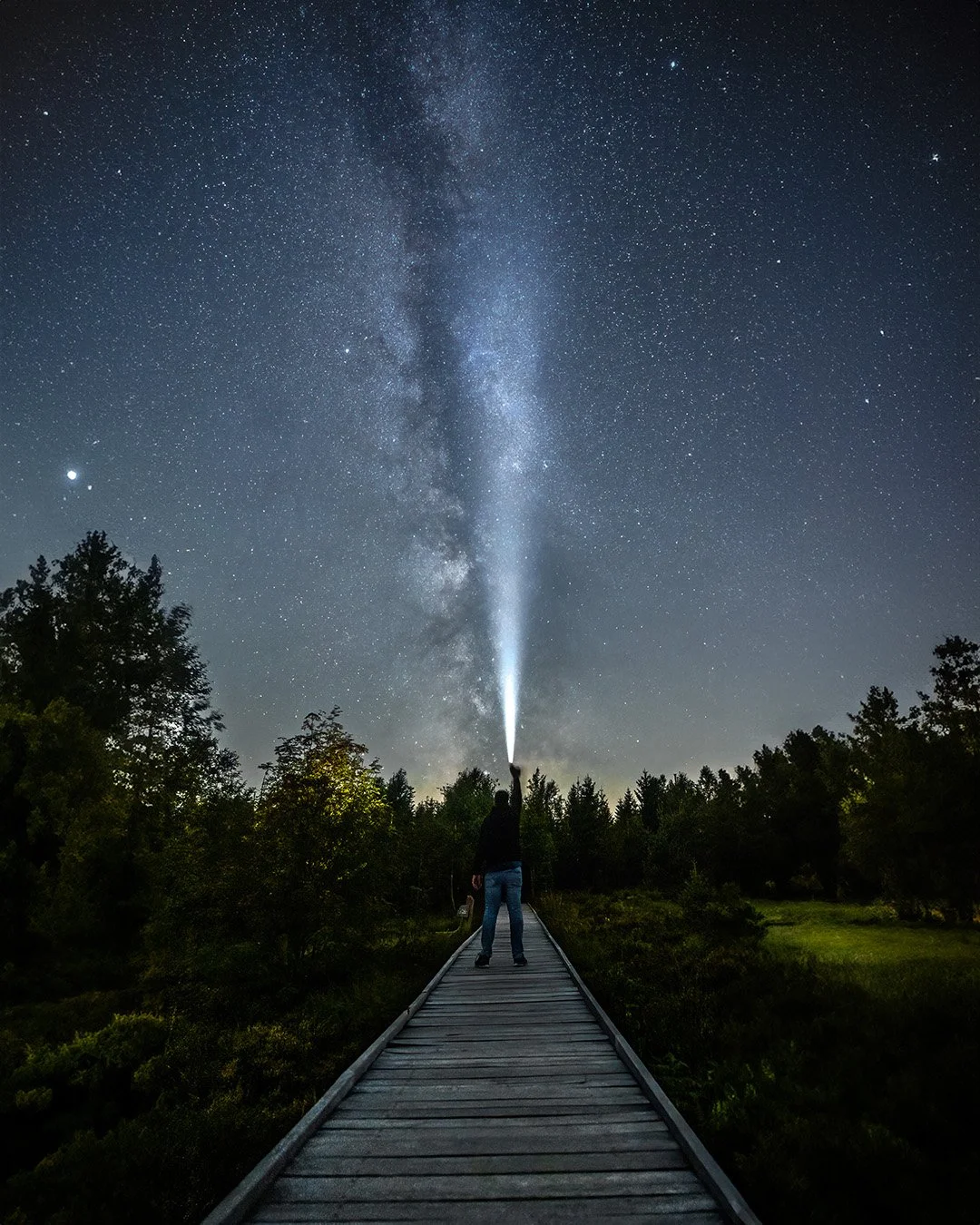 Person steht auf einem Holzsteg im Dunkeln, richtet eine Taschenlampe in den Himmel mit Sternen und der Milchstraße