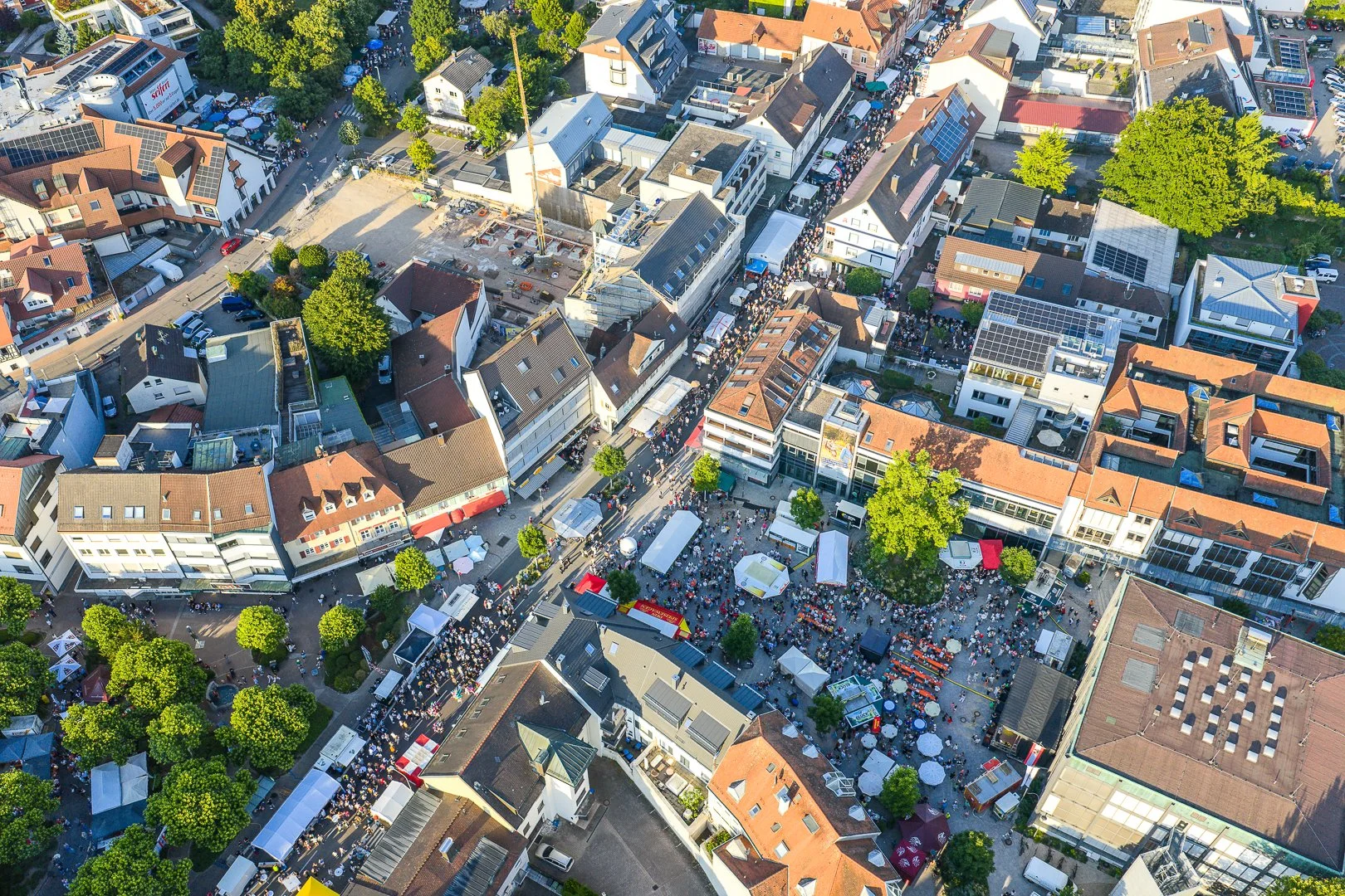 Luftaufnahme einer belebten Innenstadt während eines Festivals mit Ständen, Zelten, Menschenmengen, Bäumen und Gebäuden.