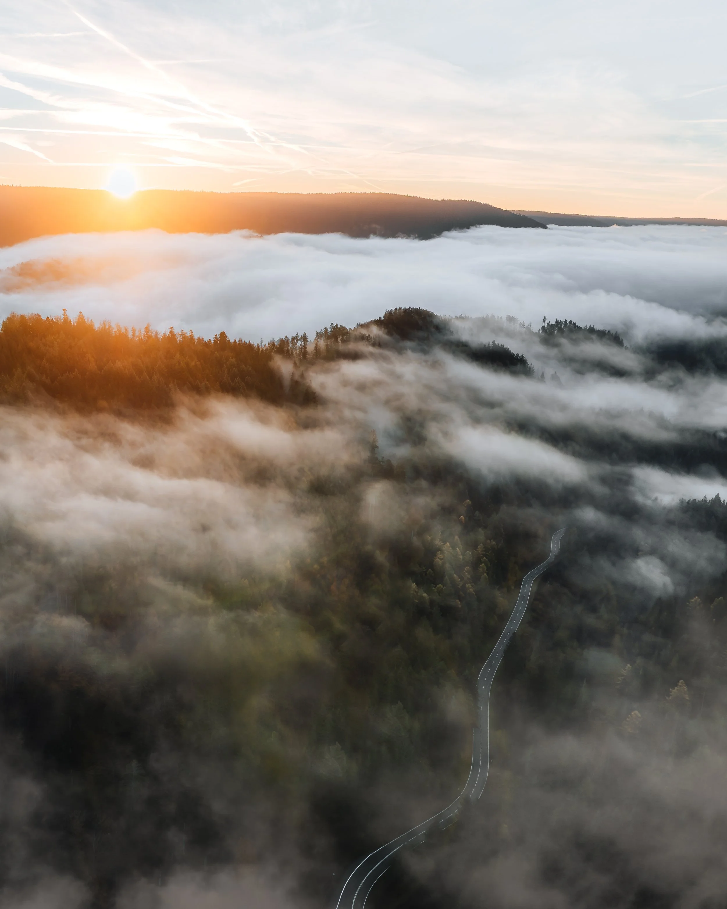 Vogelperspektive auf eine bewaldete Gebirgsregion bei Sonnenaufgang, mit Wolken und Nebel, einer kleinen kurvigen Straße im Tal.