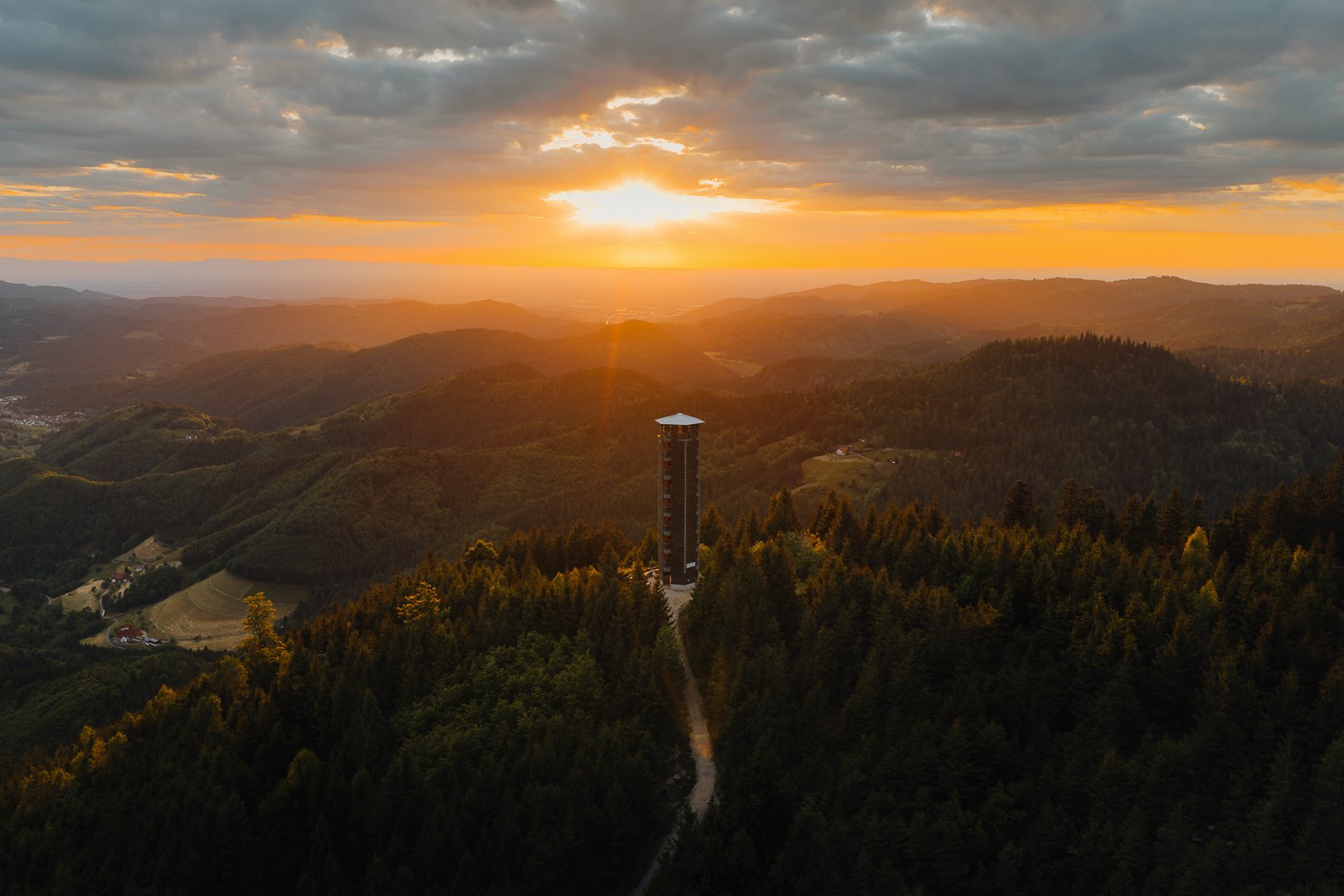 Berglandschaft bei Sonnenuntergang mit einem hohen Turm in der Mitte, umgeben von Wäldern.