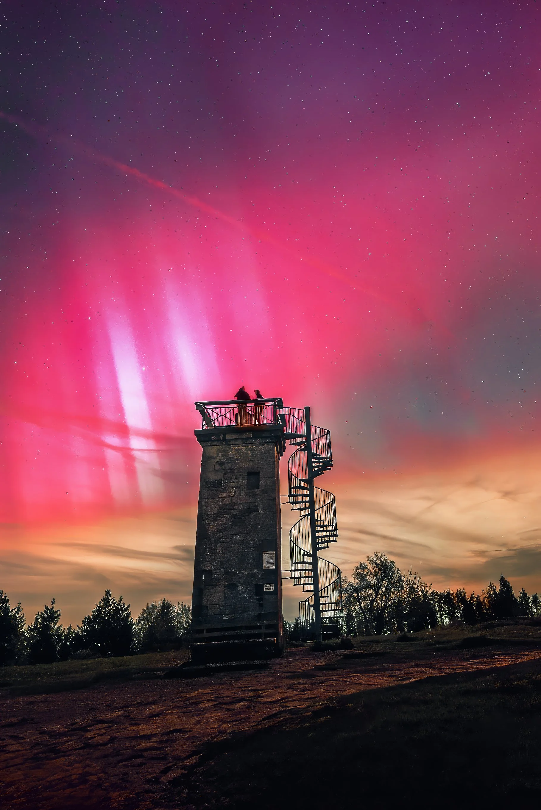 A lighthouse with a spiral staircase stands in front of the colorful aurora borealis and a starry sky.