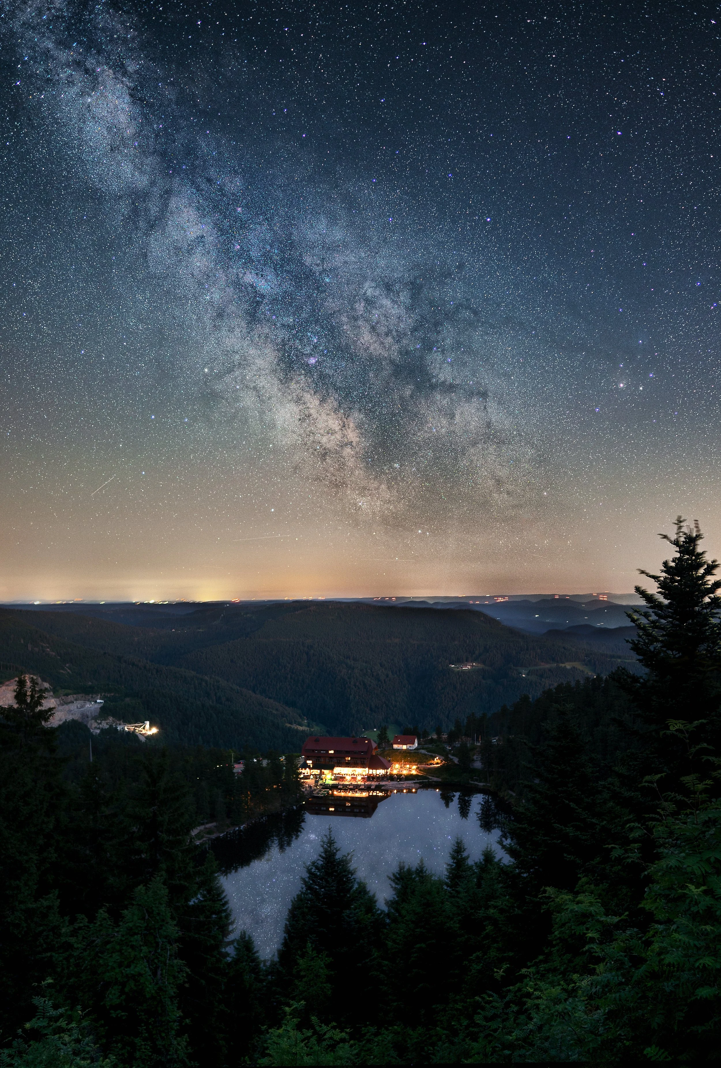 Sternenbedeckter Nachthimmel über einer ruhigen Bergregion mit einem kleinen See, umgeben von Tannen, und einem Haus mit Licht im Vordergrund, das den Himmel spiegelt.