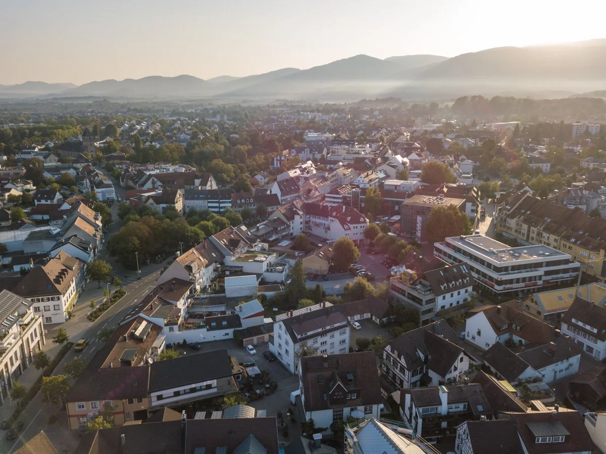 Luftaufnahme einer Stadt mit Häusern, Straßen, Bäumen und Hügeln im Hintergrund bei Sonnenuntergang.