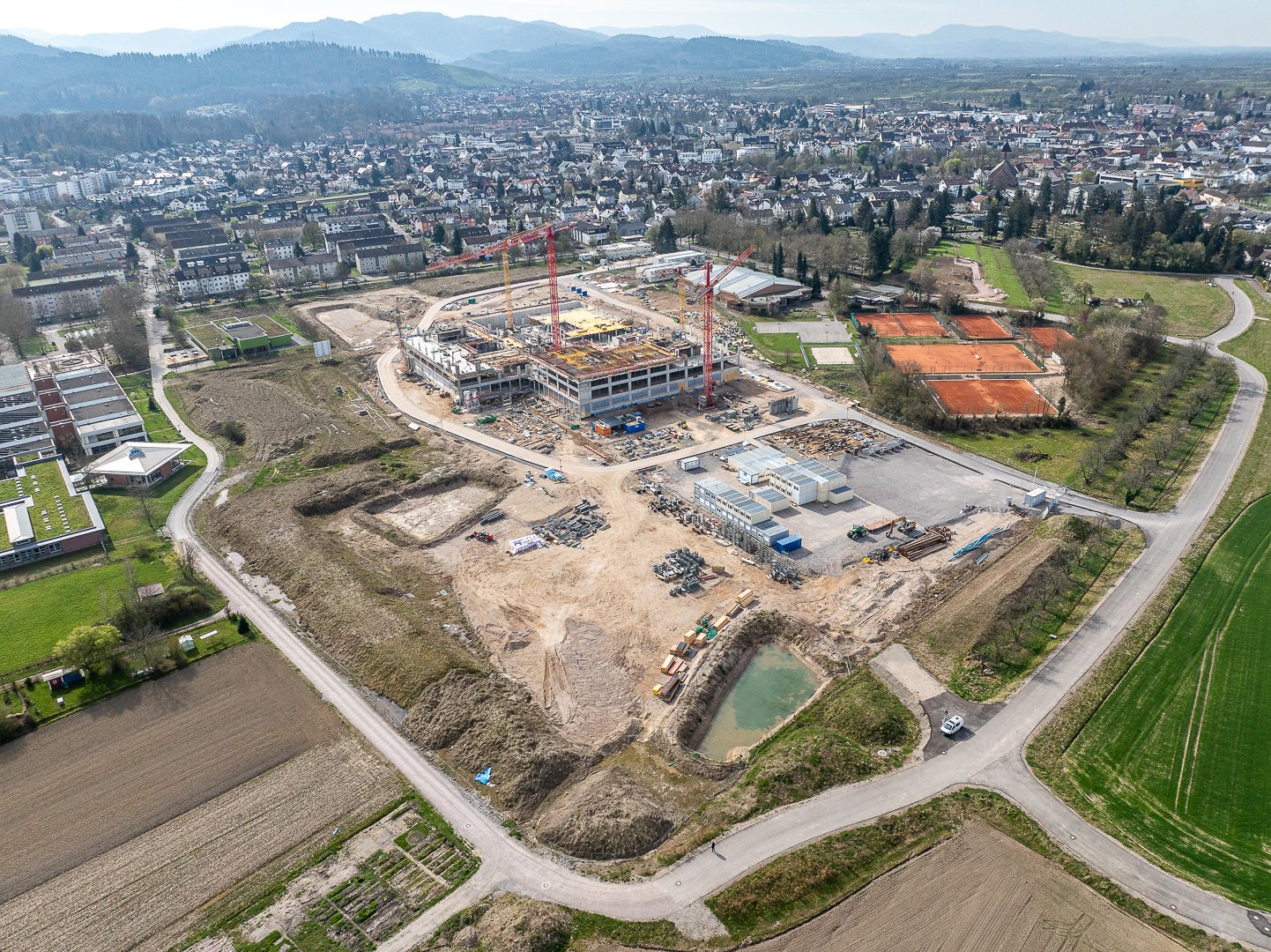 Baufeld mit kranbegleitetem Mehrfamilienhausbau, umgeben von Feldern, einem kleinen See und einer Stadt im Hintergrund.