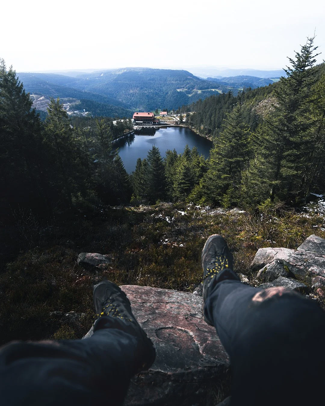 Bergsteiger sitzt auf einem Felsen und blickt auf einen See, umgeben von dichten Wäldern und Bergen, mit einer Hütte am Ufer.