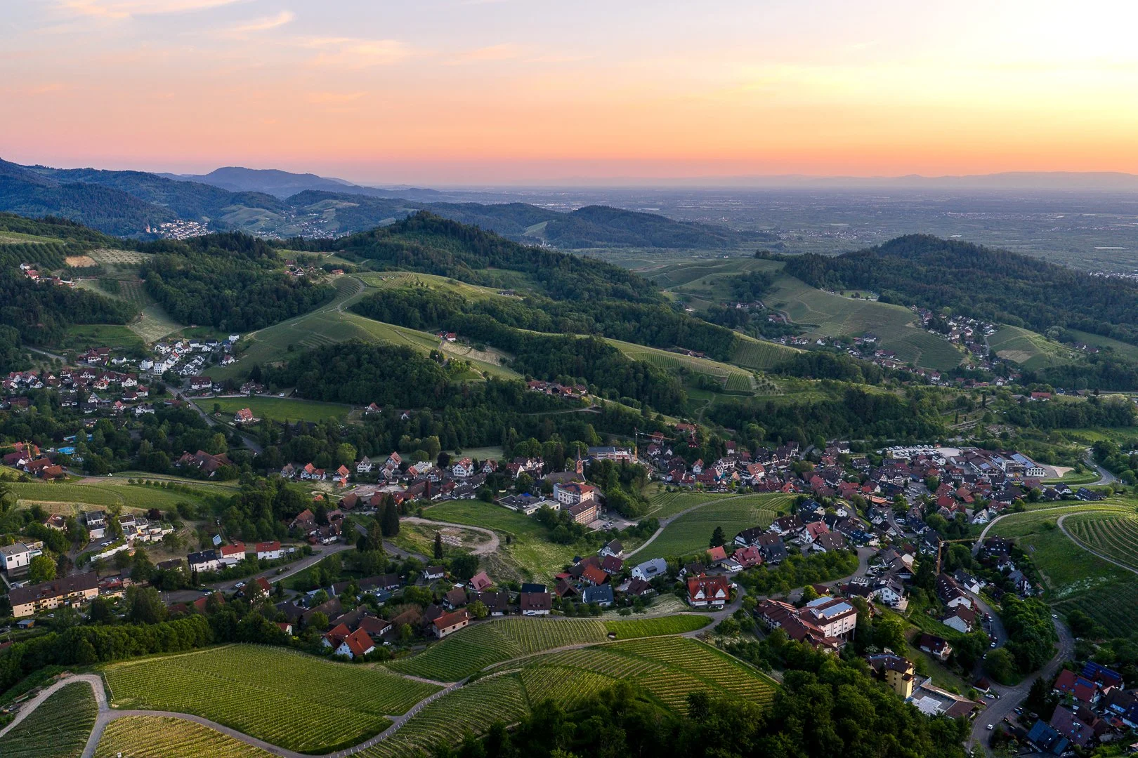 Landschaftliches Bild von Hügeln, Wäldern, Weingärten und einem Dorf bei Sonnenuntergang.