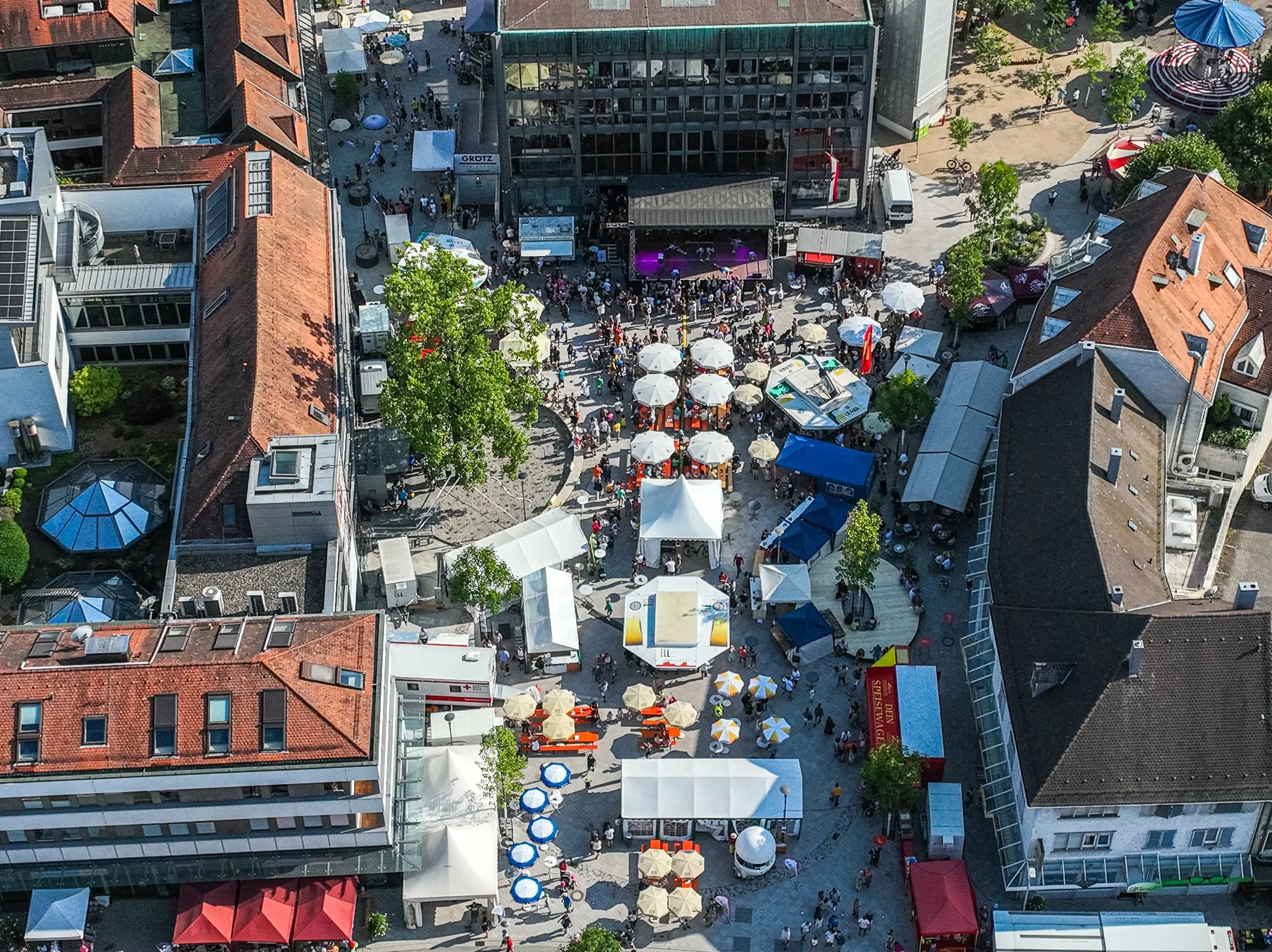 Luftaufnahme eines belebten Stadtplatzes mit Ständen, Zelten und Personen bei einem Fest oder Markt in der Mitte. Es gibt Tische mit Sonnenschirmen, Bäume und umgebende Gebäude mit roten und grauen Dächern.
