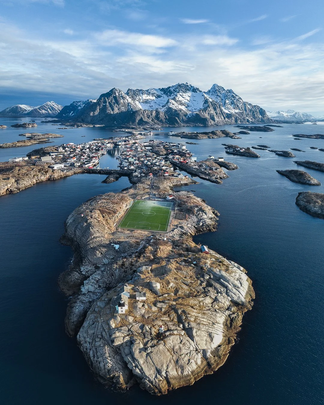 Drohnenaufnahme einer kleinen Insel vor den Bergen mit einem Fußballfeld, umgeben von Wasser und kleinen Häusern.