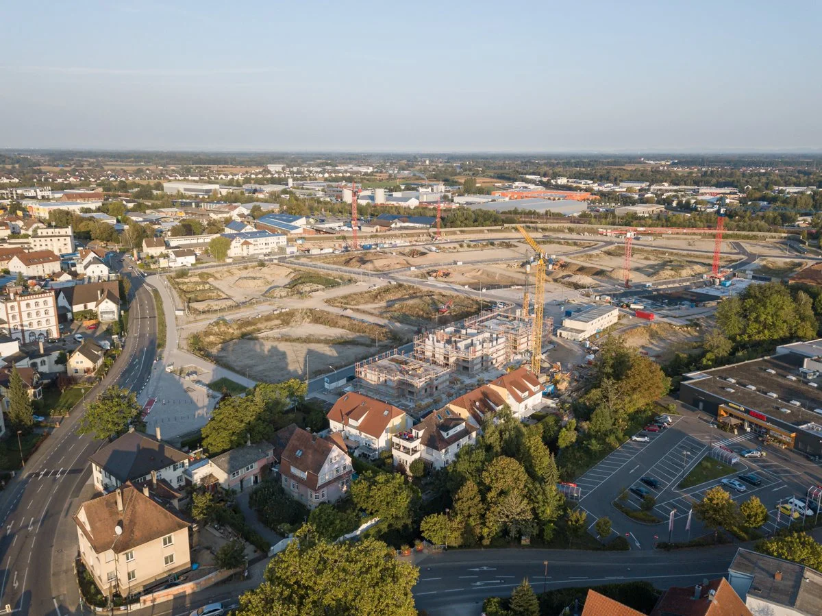Luftaufnahme einer Stadt mit Bauarbeiten, Baukränen und Wohnhäusern im Vordergrund, sowie Industrie- und Gewerbegebäude im Hintergrund.