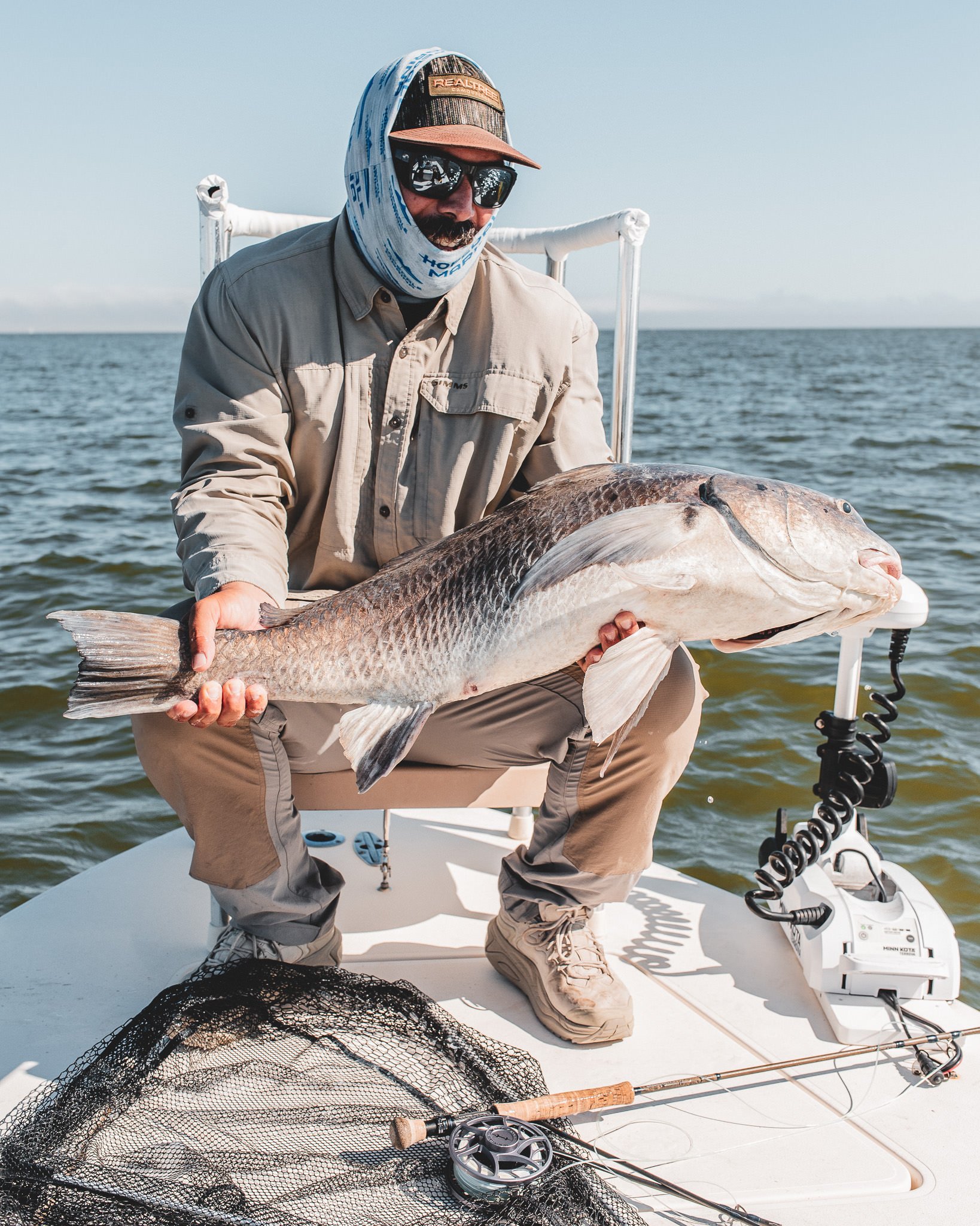 A man in fishing attire, wearing a hat, sunglasses, and a neck gaiter, holds a large fish on a boat with fishing gear and a net, with water in the background.
