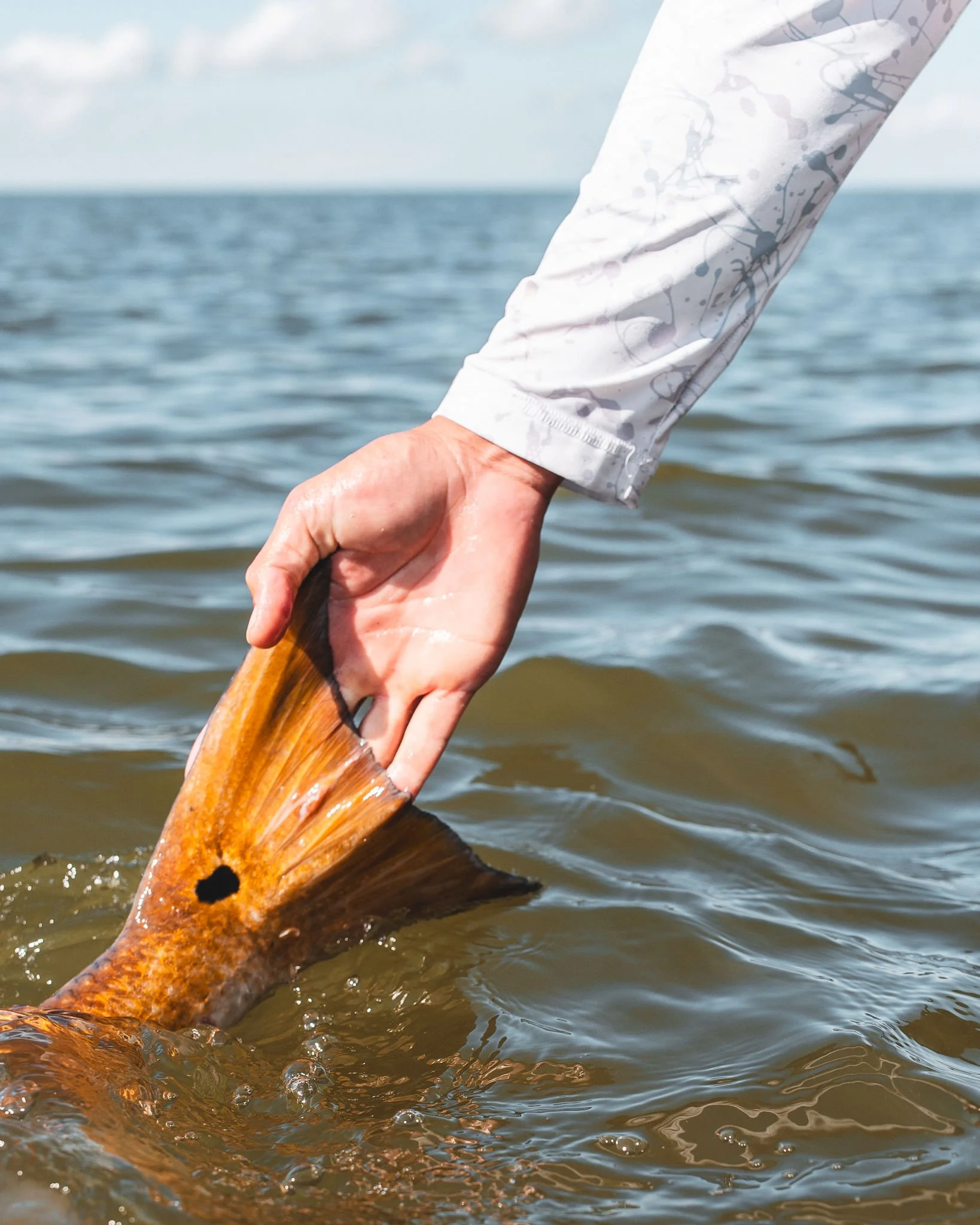 A hand holding a fish tail in water, with a person's sleeve showing a light-colored, patterned shirt, and a body of water in the background under a partly cloudy sky.