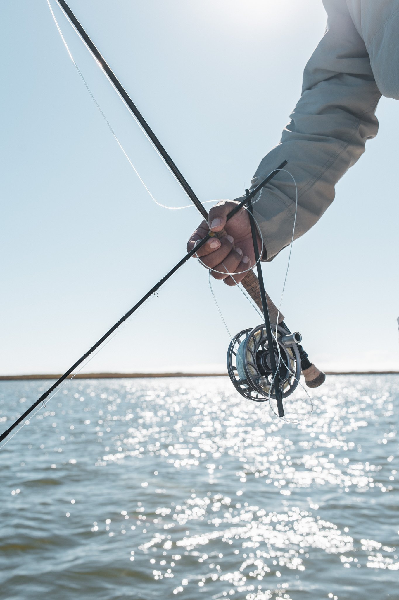 A person holding a fishing rod and reel with flies attached, standing near a body of water with the sun shining, in a bright outdoor setting.