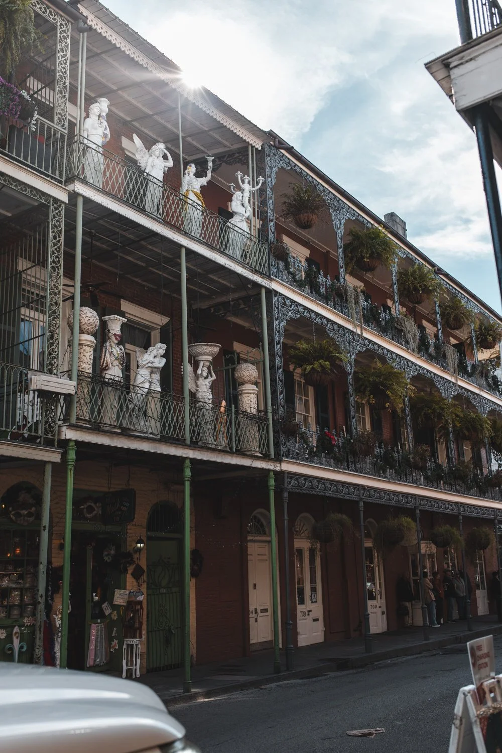 Three-story building with ornate iron balconies decorated with statues, plants, and architectural elements, under a partly cloudy sky with sunlight.