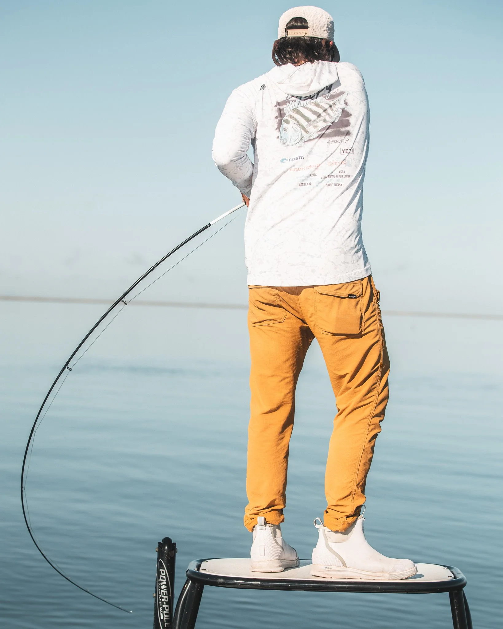 A person standing on a small platform fishing in a body of water, wearing khaki pants, a white hoodie with fish design, a gray cap, and white sneakers, with a fishing rod in hand.