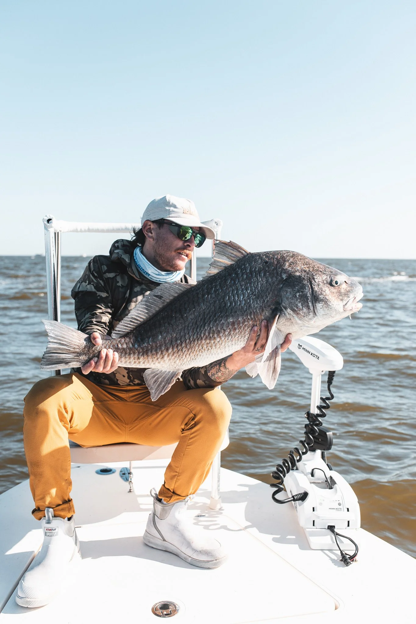 Man on a boat holding a large fish, wearing sunglasses, a light-colored cap, a camouflage jacket, tan pants, and white boots, with water and sky in the background.