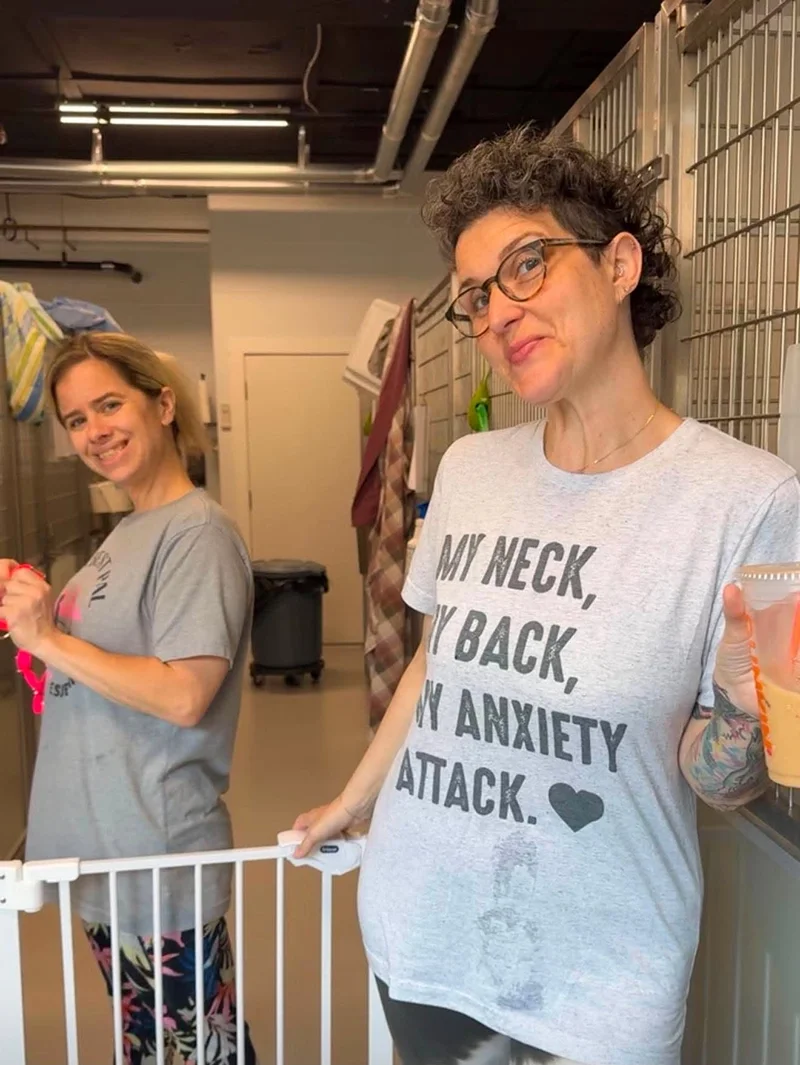 Two women standing behind a white pet gate in an indoor setting, smiling at the camera. One woman is wearing glasses and a gray shirt with text about anxiety, the other woman is in a gray shirt and colorful leggings.