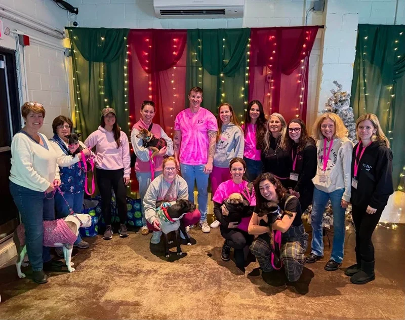 Group of women and dogs posing indoors in front of holiday-themed backdrop.