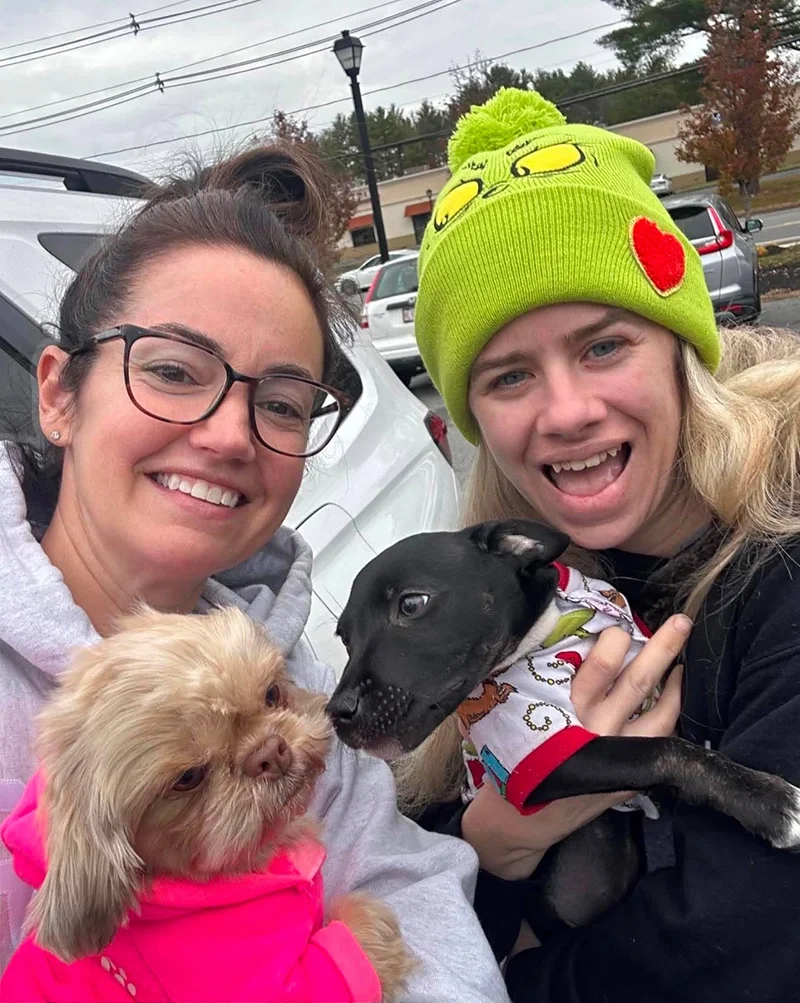 Two smiling women in casual clothes holding two small dogs, one with light fur in a pink hoodie and the other with black fur and a colorful bandana, outdoors in a parking lot with cars and trees in the background, one woman wearing a bright green knit hat with a red heart and cartoon face design.