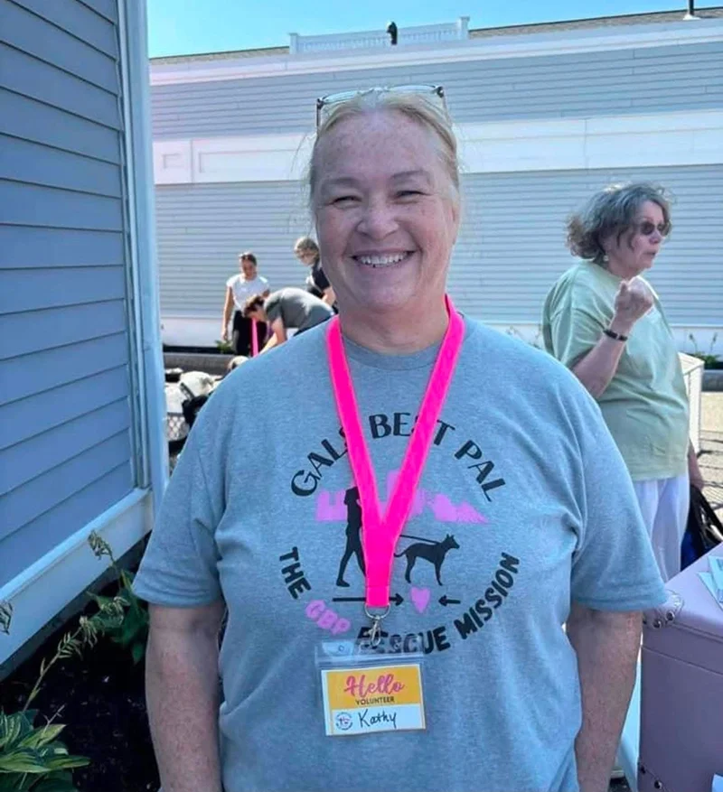 Woman smiling at a volunteer event, wearing a gray T-shirt with a Gals Best Pal logo and a bright pink lanyard with a volunteer badge labeled 'Kathy.'