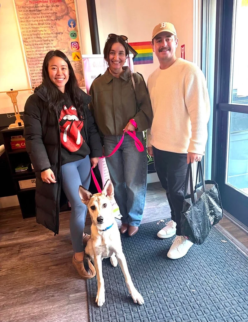 Three people standing inside a building with a dog sitting on the floor in front of them. The woman on the left is wearing a black jacket and a hoodie with a tongue and lips graphic. The woman in the middle is holding a pink leash attached to the dog