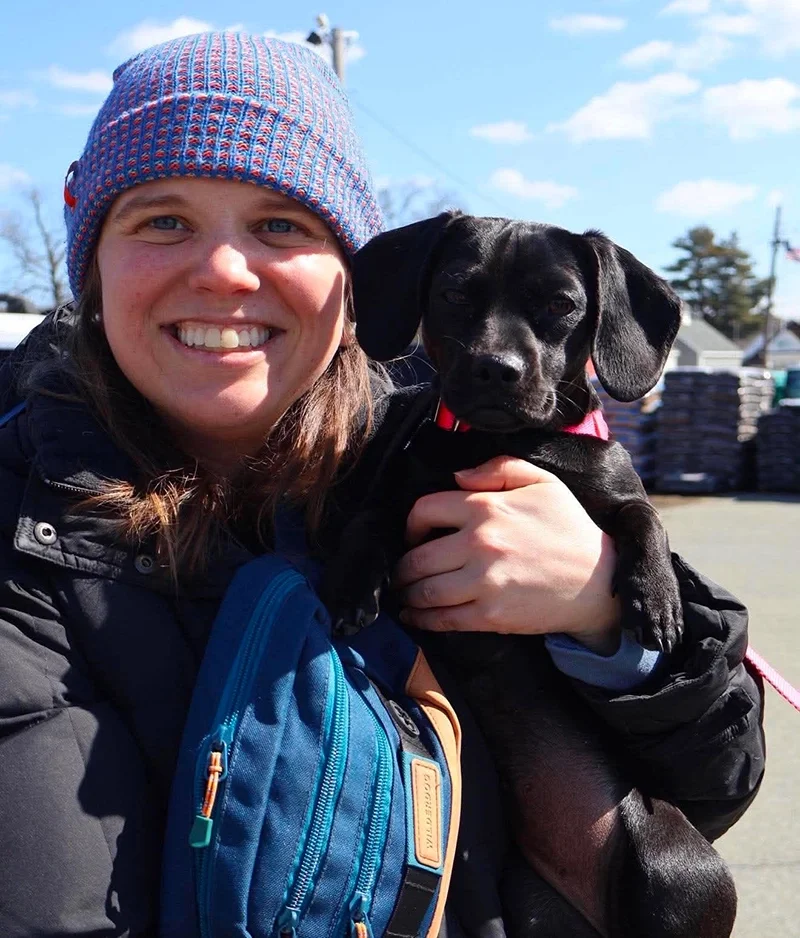 Smiling woman in a colorful knit beanie holding a small black dog outdoors on a sunny day.