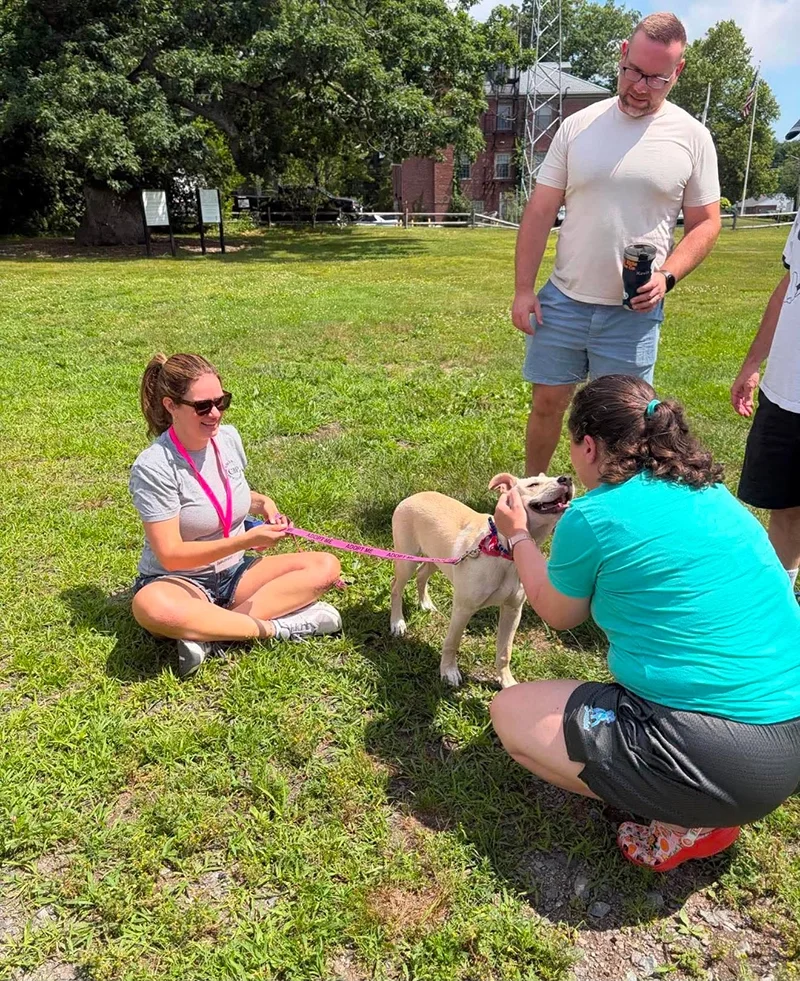 A group of four people and a dog are outside on a grassy area. One woman is sitting on the ground holding the dog's leash, smiling at the dog. Another woman is kneeling in front of the dog, petting it. Two men stand nearby, one holding a drink cup an