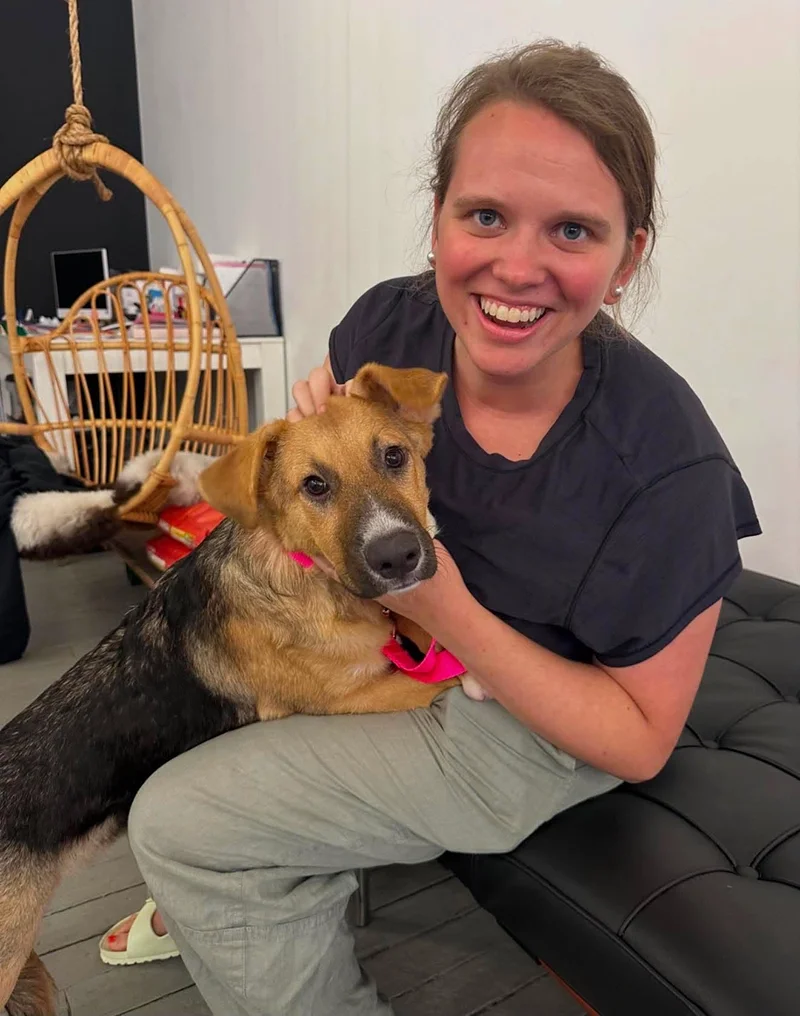 A woman sitting on a black bench, smiling at the camera, with a dog in her lap. The dog has a brown and black coat with a pink collar. The background features a white wall, a hanging wicker chair, and some electronic devices.