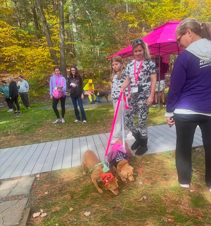 People at an outdoor Gals Best Pal event with dogs on a wooded trail, some under a pink canopy, autumn foliage in the background.