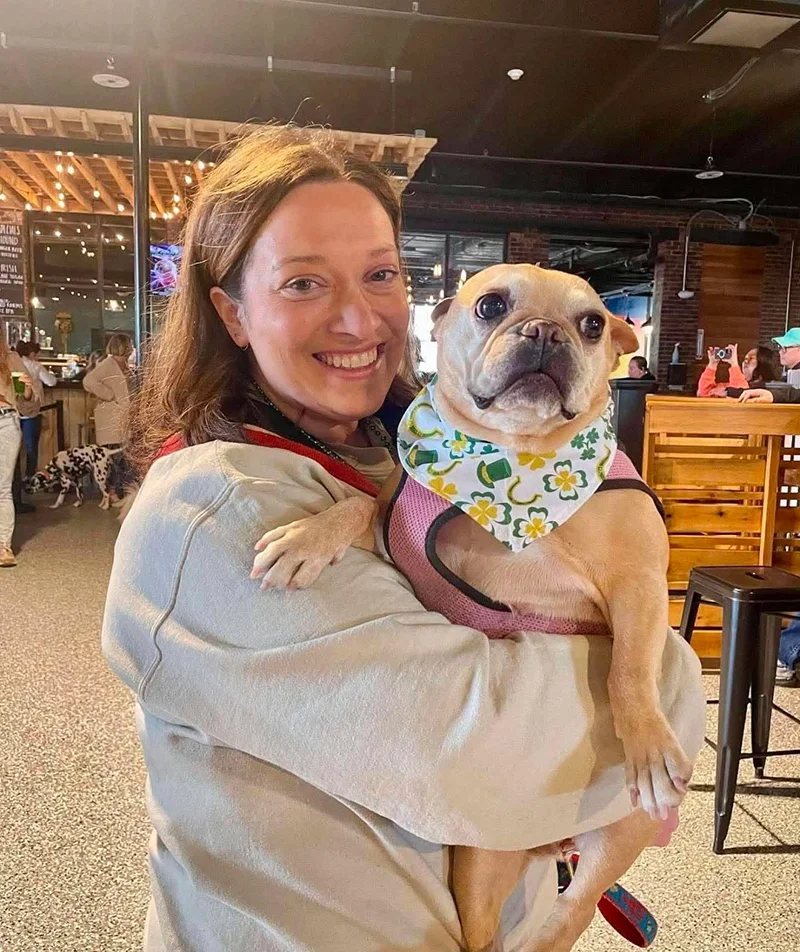 A woman smiling and holding a dog wearing a colorful bandana inside a lively indoor venue.