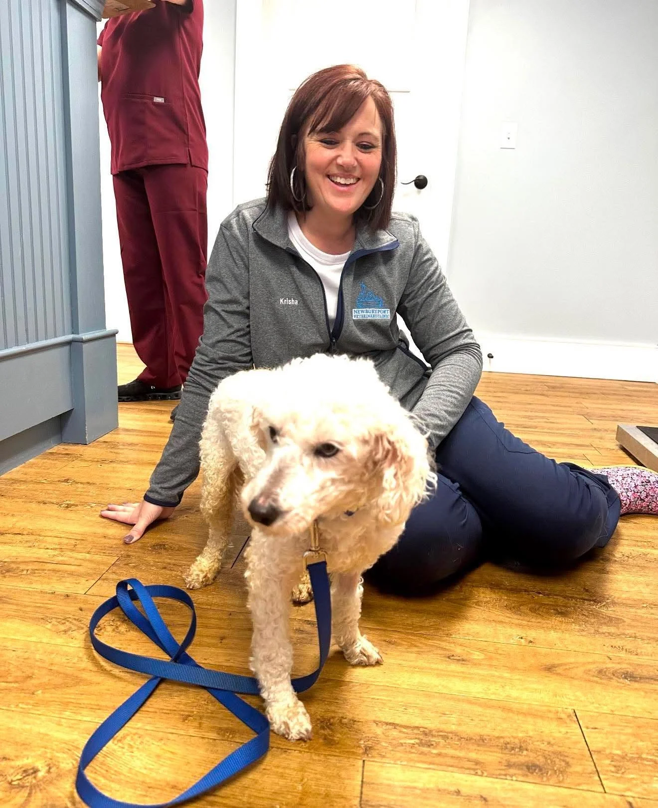 A woman sitting on the wooden floor with a happy expression, holding a puppy on a leash. She is indoors with a white wall, a door, and part of a person in red scrubs in the background.