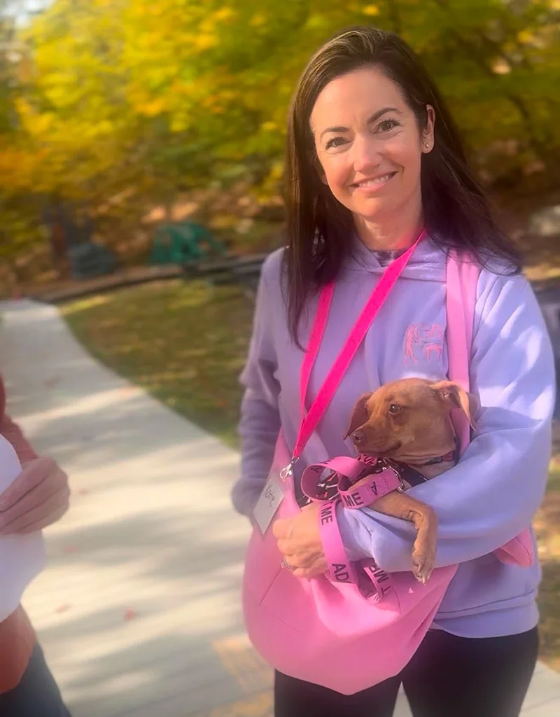 A woman smiling outdoors holding a small brown dog in her arms, wearing a lavender sweatshirt and pink bag, with trees and a pathway in the background.