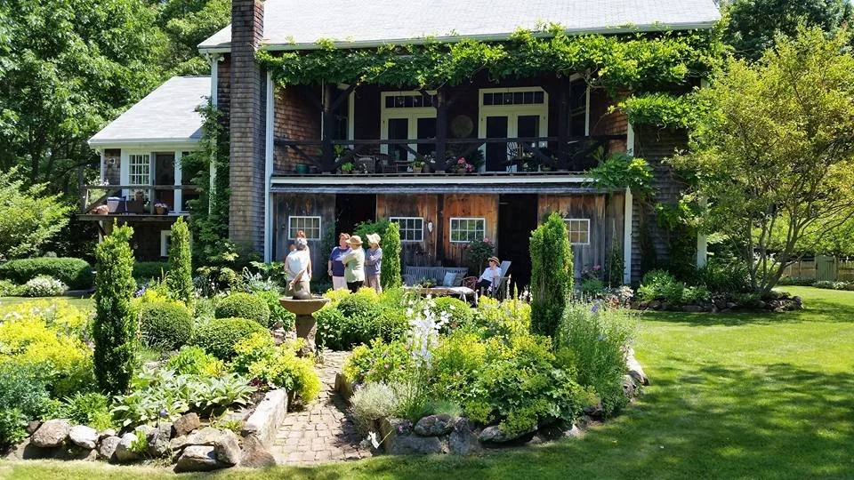 Group of people gathered in a lush garden in front of a rustic two-story house with a balcony and greenery-covered roof, surrounded by vibrant flowers and trees.