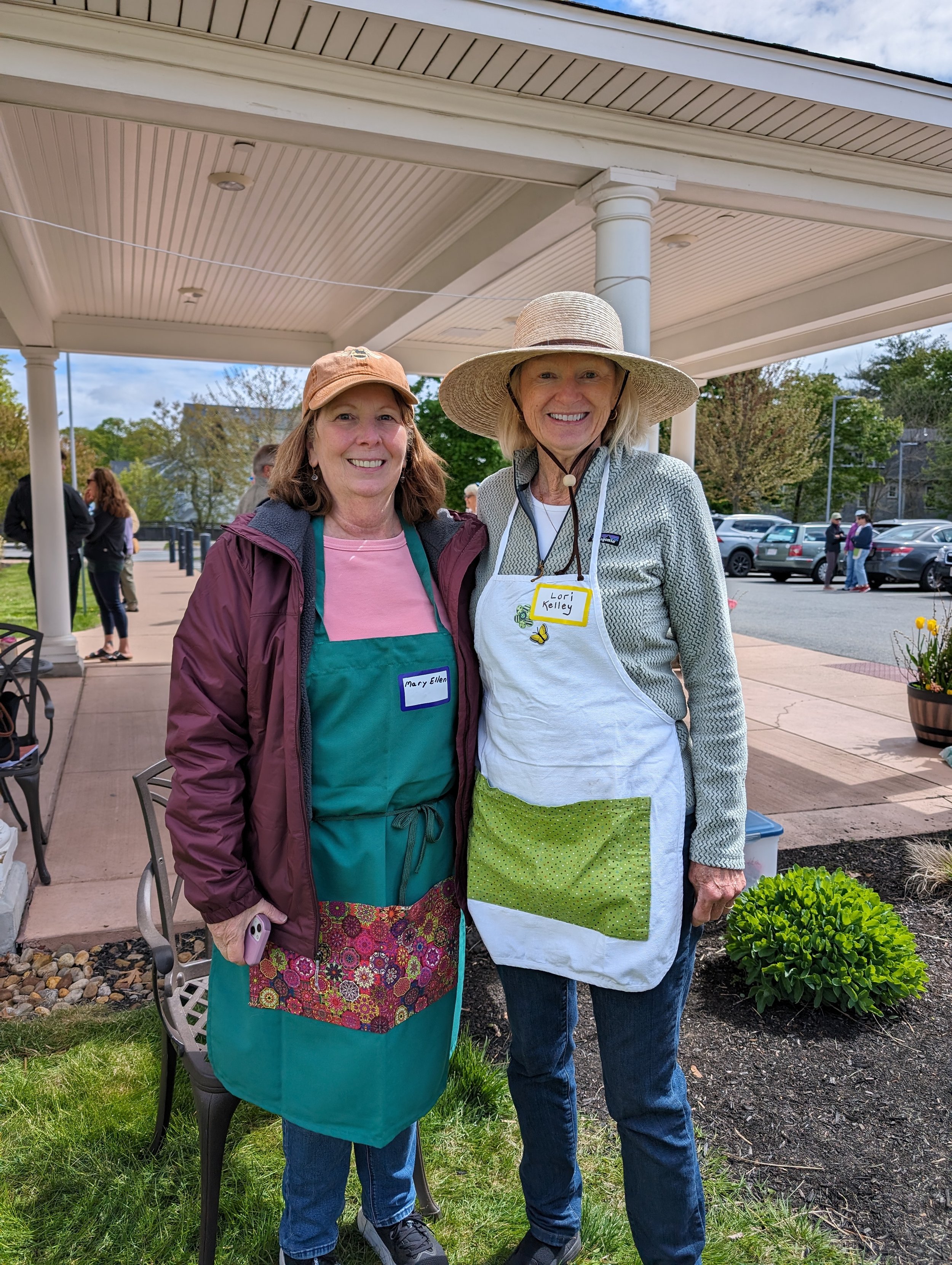 Two women standing outdoors under a porch roof, smiling for the camera. One woman wears a tan hat and apron with green polka dot pocket, and the other wears a large straw hat and name badge that reads 'Lori Kelley'. In the background, other people are socializing, and there are cars parked near trees and a building.