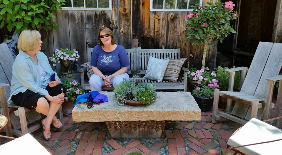 Two women sitting and talking in a backyard patio with wooden furniture, potted plants, and brick flooring.