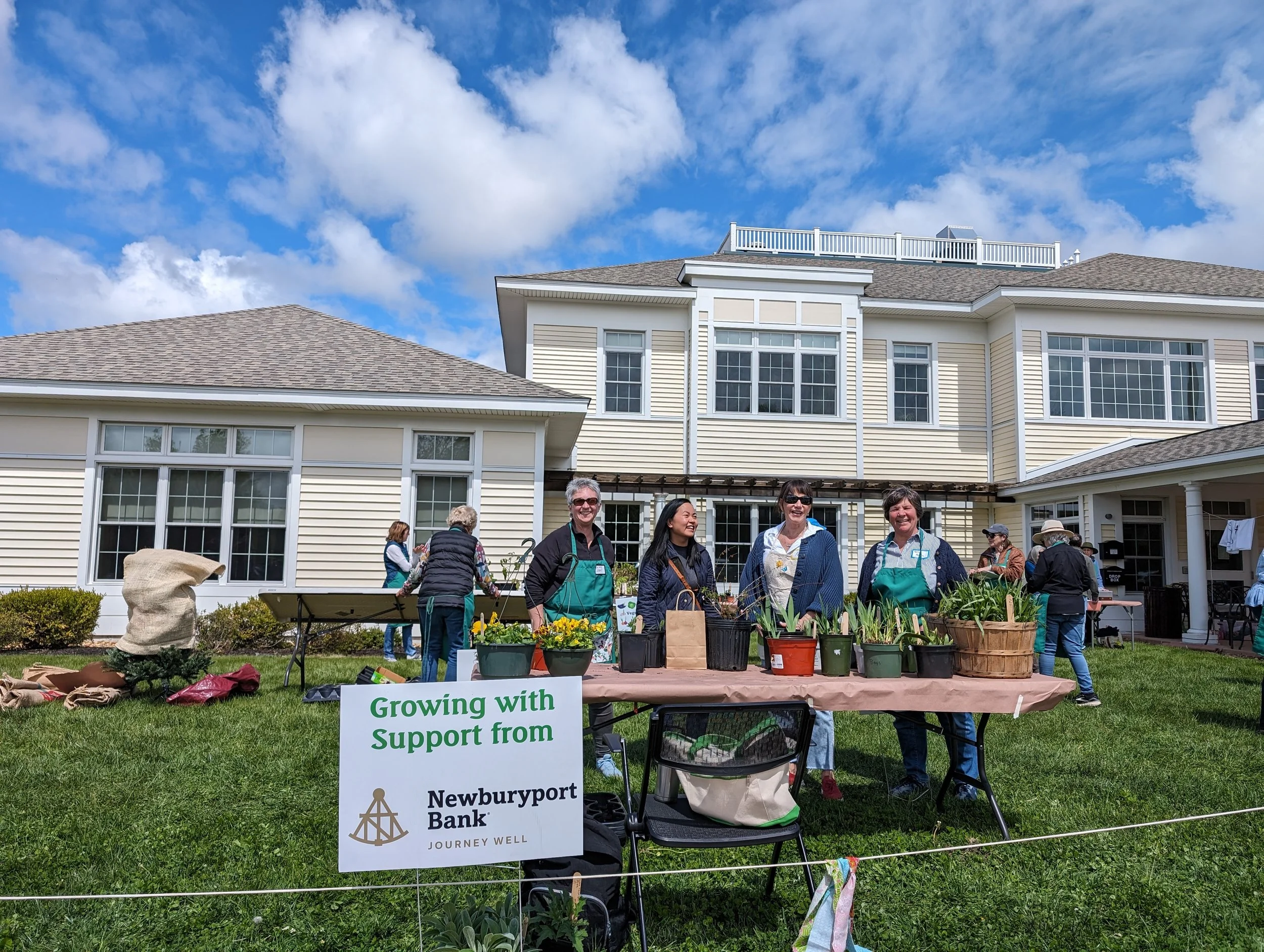 Community gardening event with people smiling, manning a table of potted plants, and a sign reading "Growing with Support from Newburyport Bank" in a backyard setting with a house and blue sky in the background.