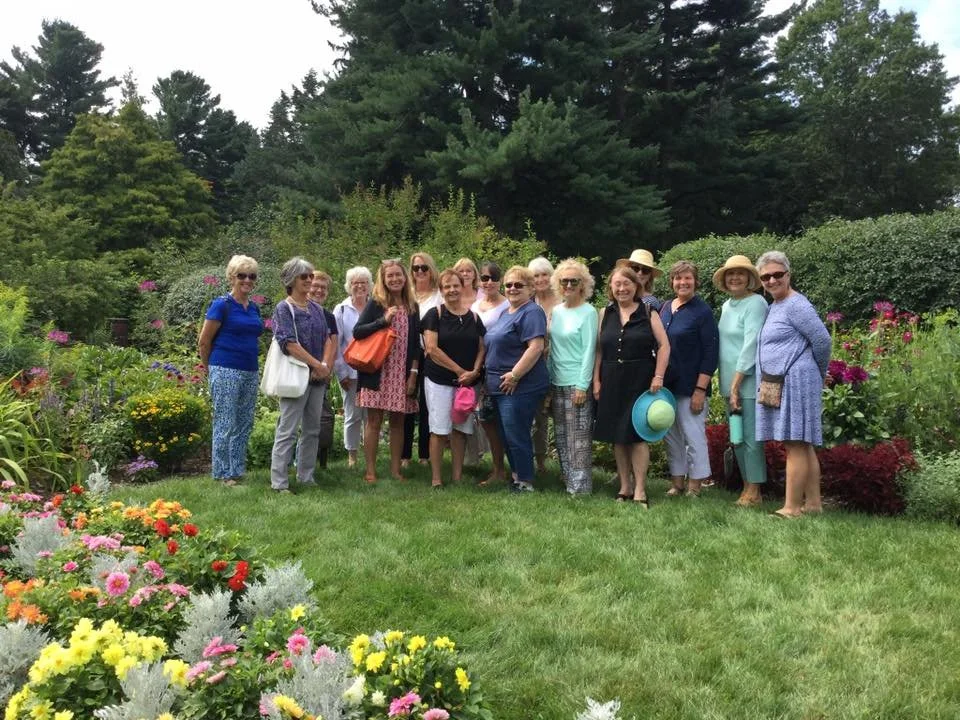 Group of women standing on a grassy lawn surrounded by colorful flowers with trees in the background.