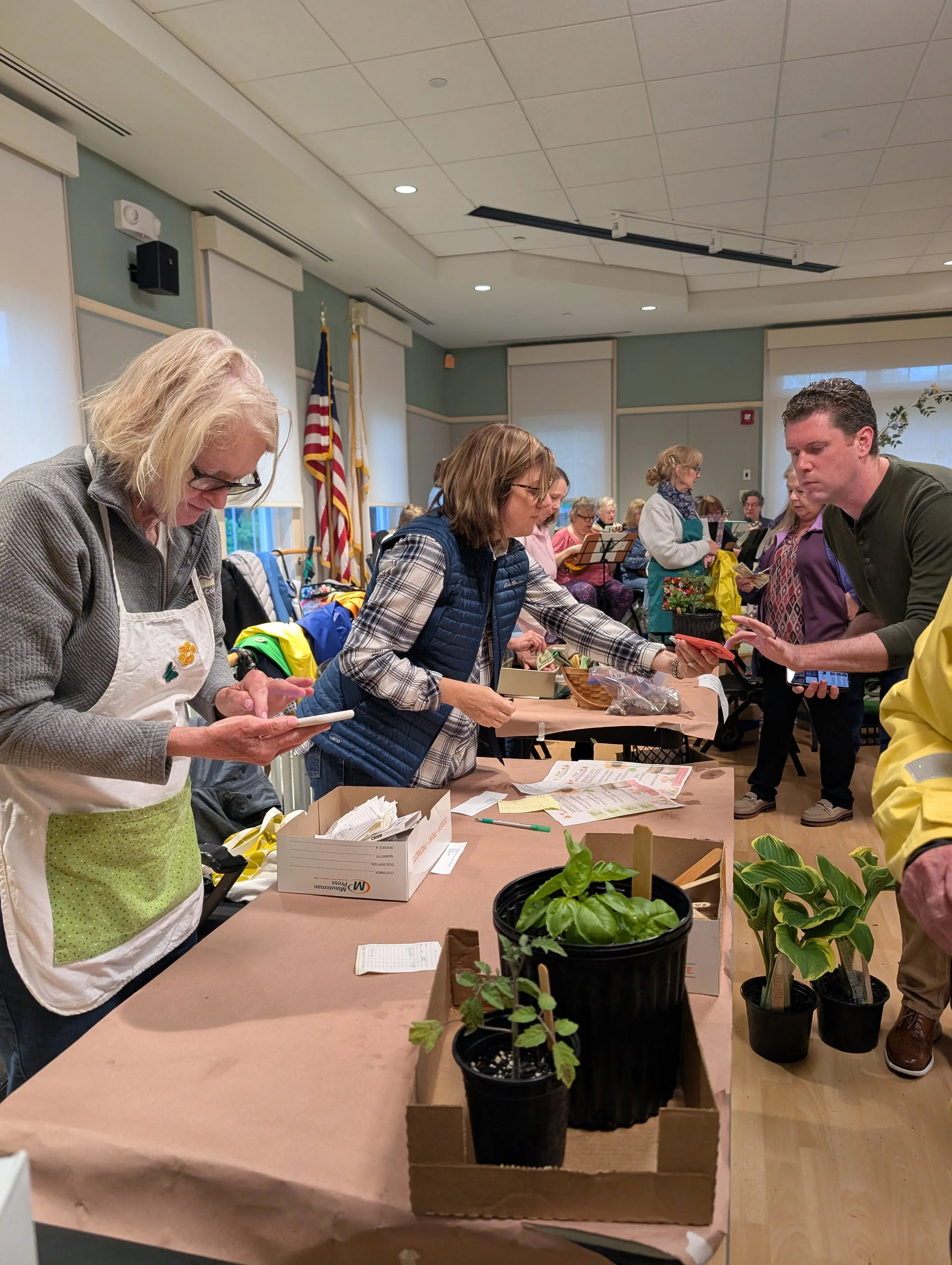 People gathered at a table with seedlings and plants, engaged in planting or selling activities in a community event or workshop, with American flags in the background.
