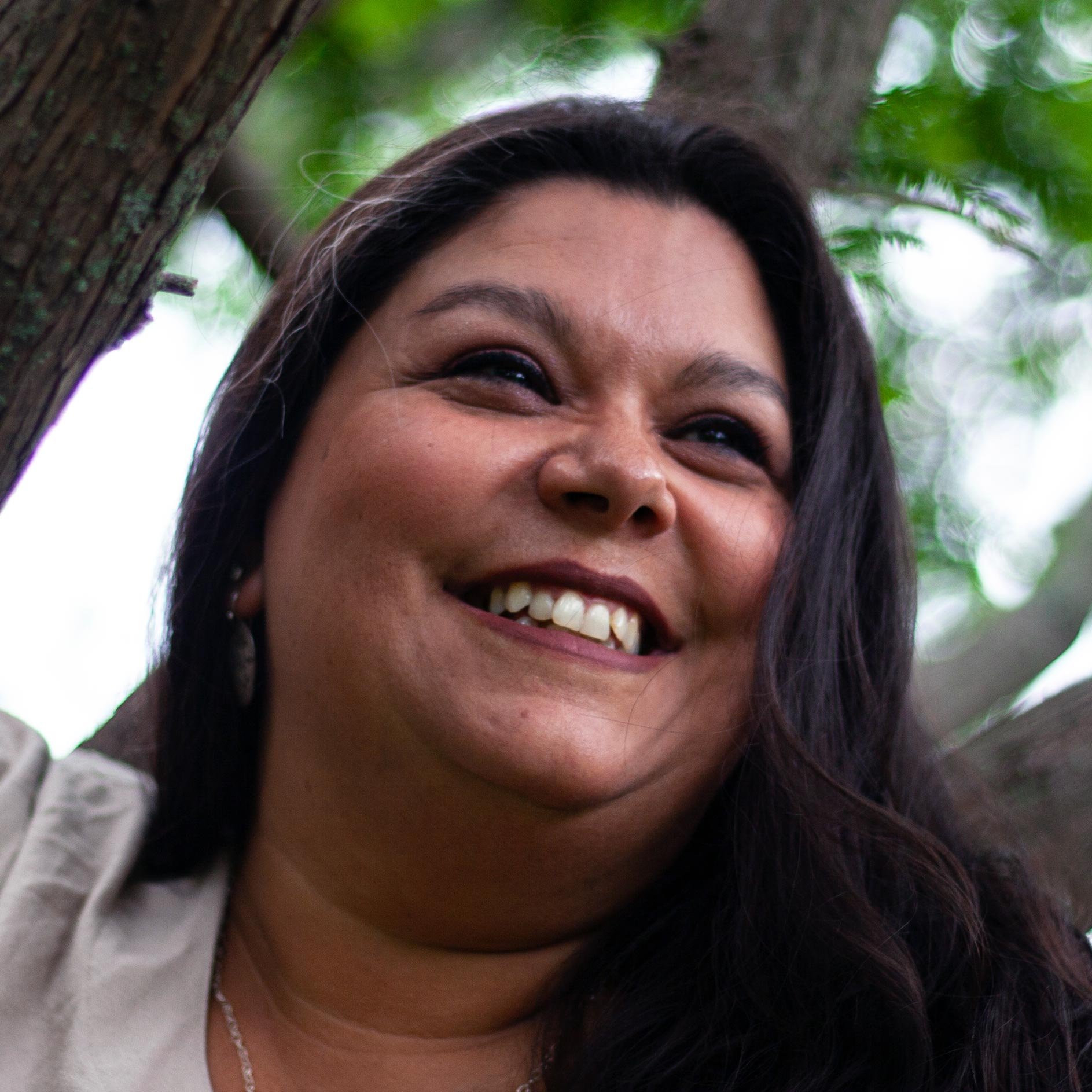 Close-up of a smiling woman with dark hair outdoors near a tree with green leaves.