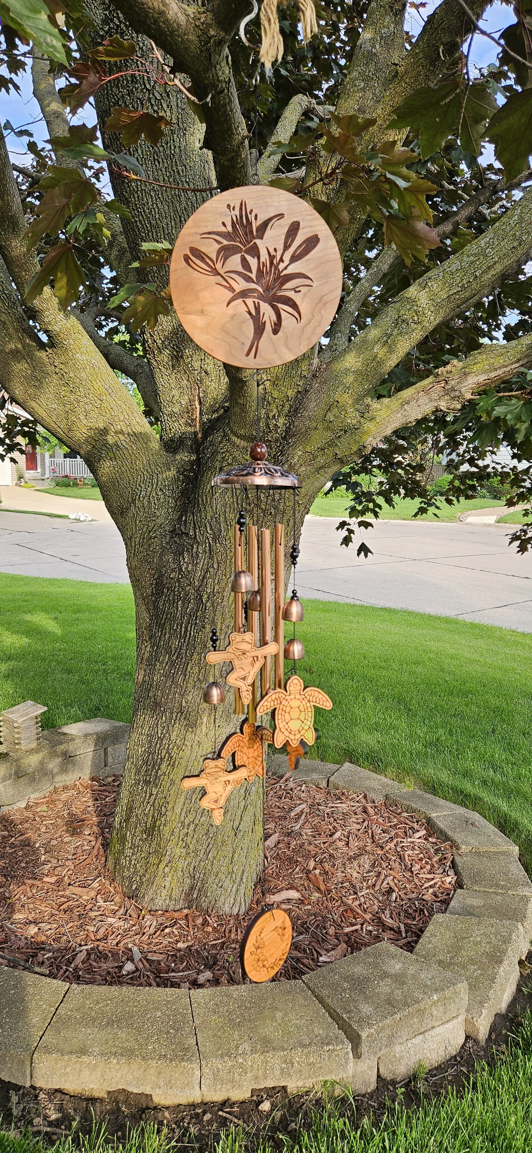 A decorative wind chime hanging from a tree branch, with a round wooden disk featuring a floral design at the top, and wooden and metal ornaments including a turtle, a fairy, and a dragonfly hanging below. The tree has a thick trunk and green leaves,
