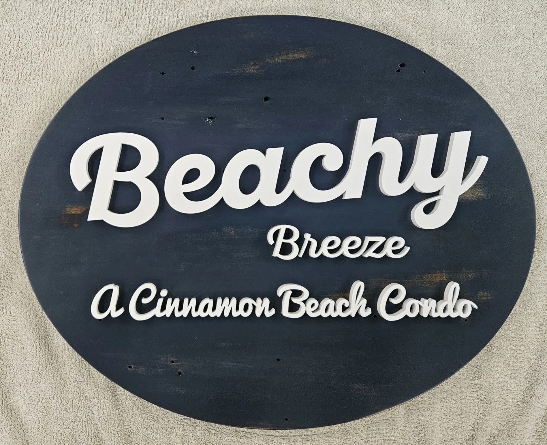 Oval sign with dark background and white raised lettering reads 'Beachy Breeze' and 'A Cinnamon Beach Condo'.
