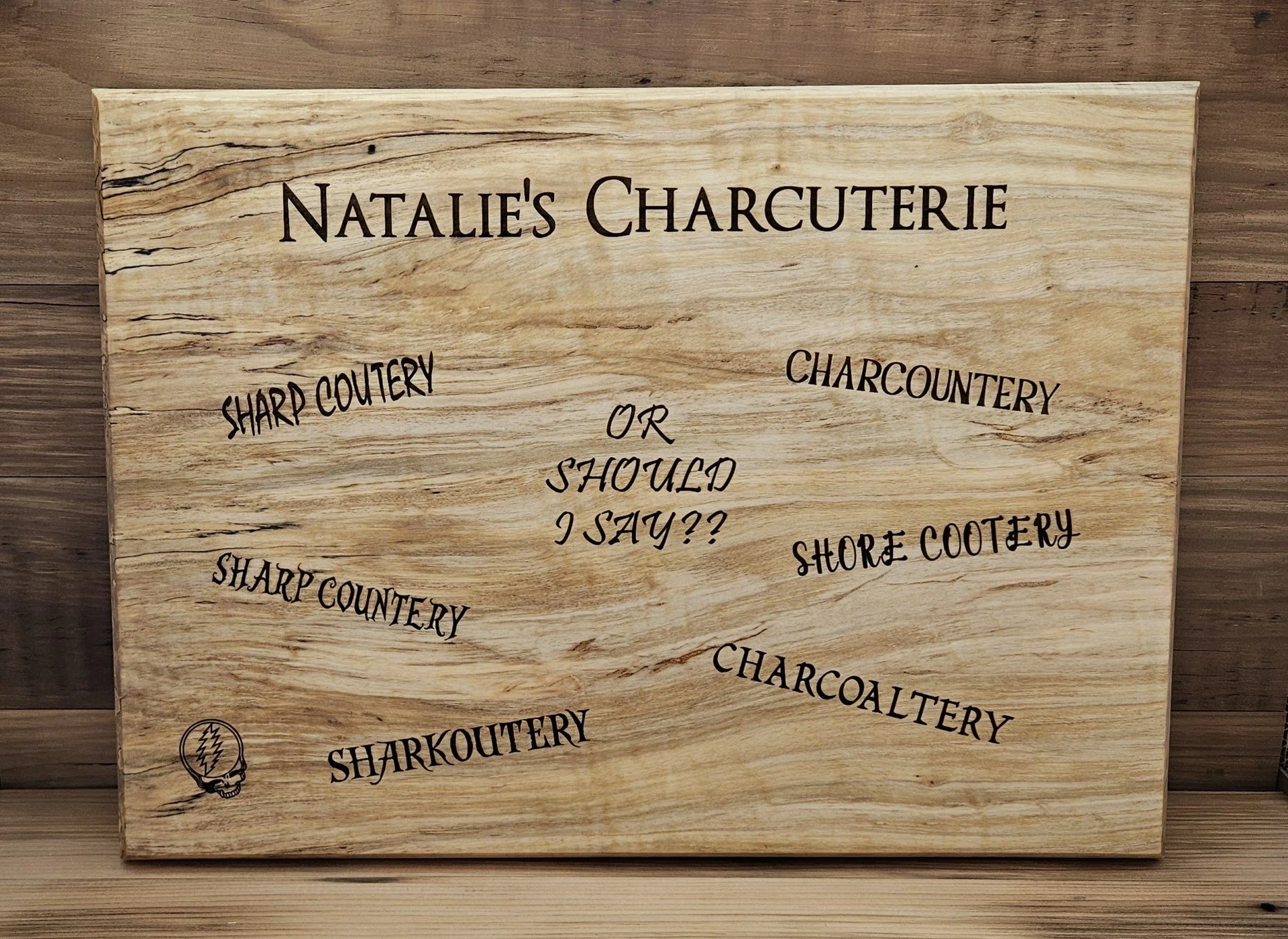 Wooden sign for Nathalie's Charcuterie with words about types of cutlery and a small skull drawing in the bottom left corner.