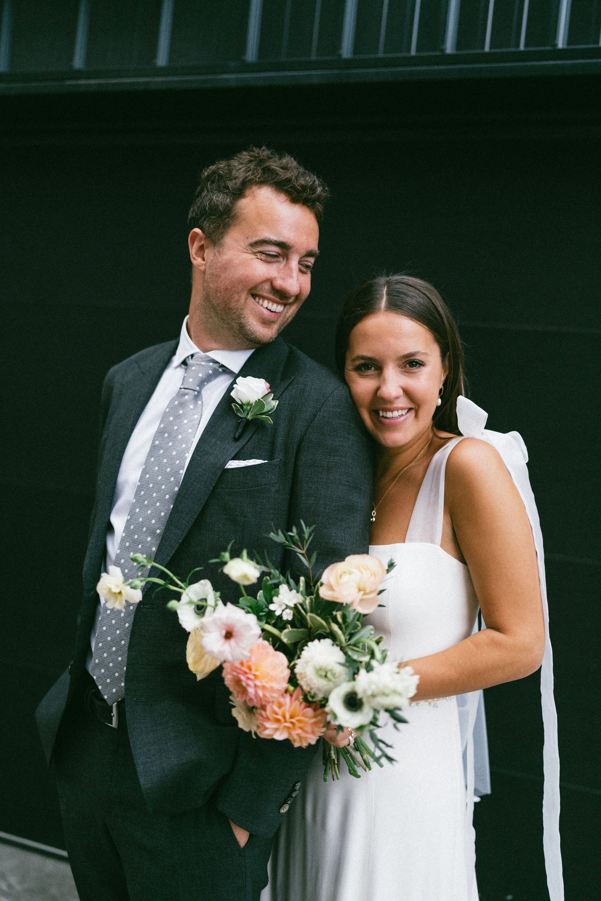 A bride and groom smiling, standing close together, with the bride holding a bouquet of flowers, against a dark background.