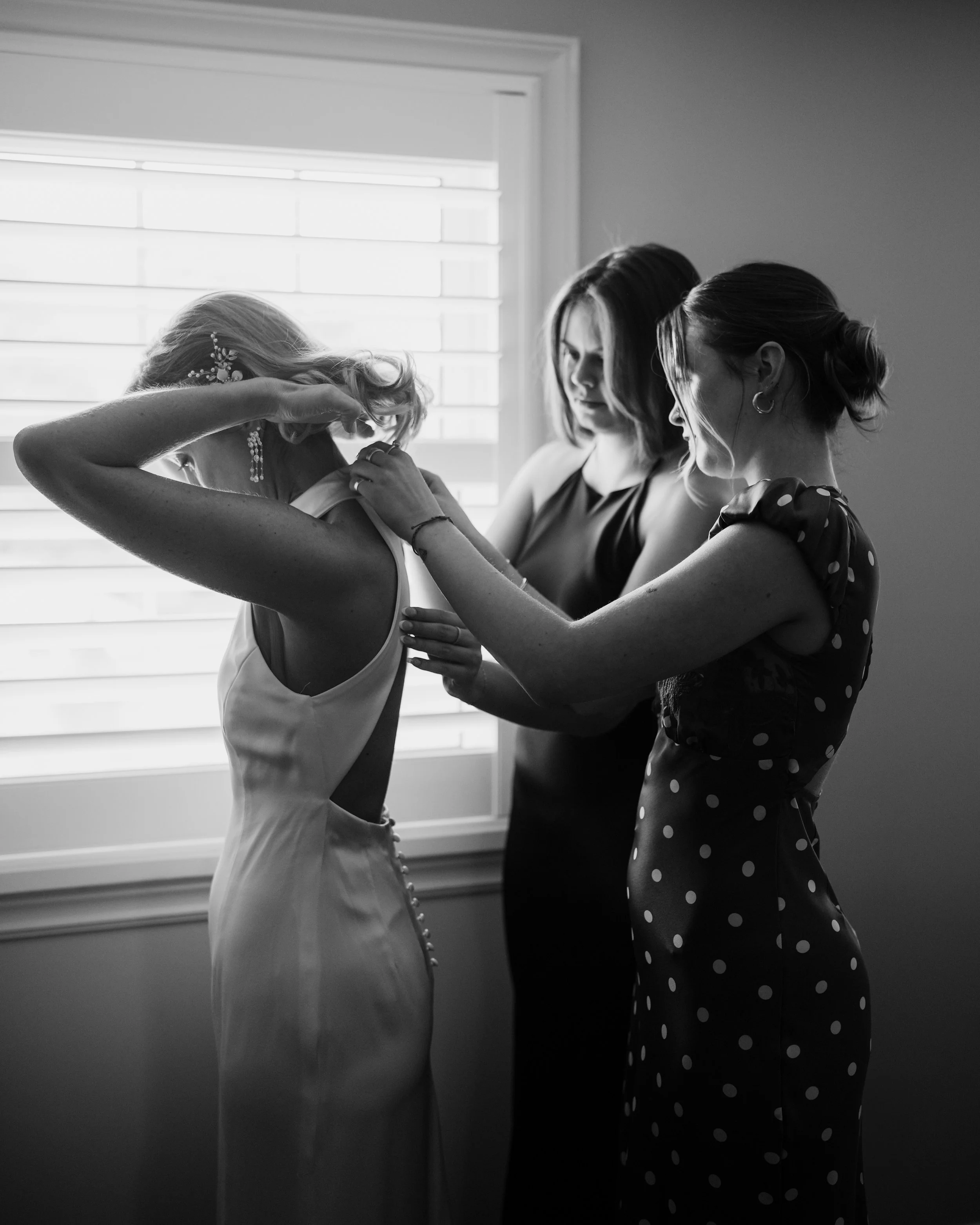 Three women preparing a bride for her wedding in front of a window with blinds, one helping with her dress and the others assisting.
