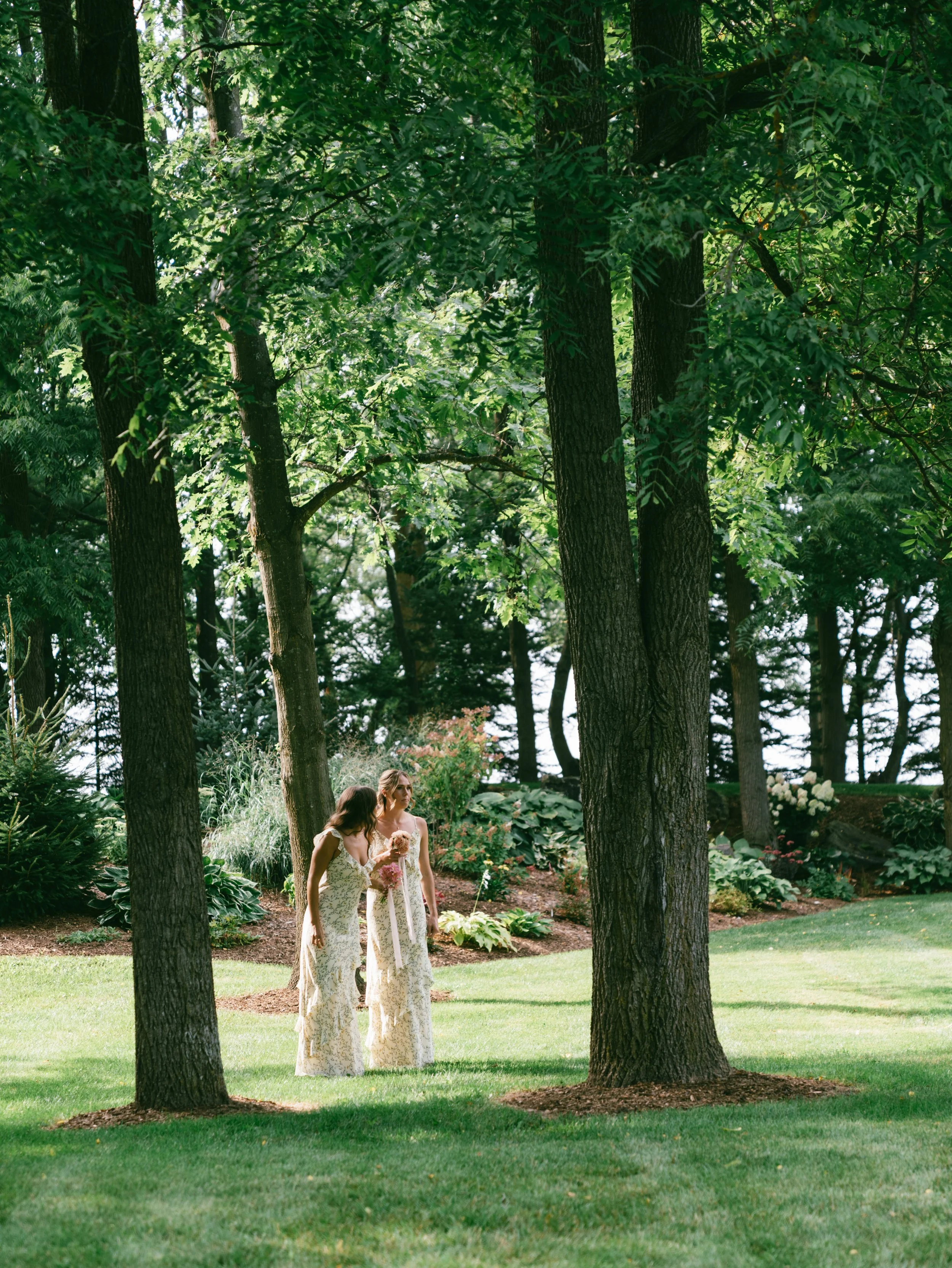 Two women in long floral dresses walking through a lush green park with large trees, shrubs, and well-maintained grassy areas.