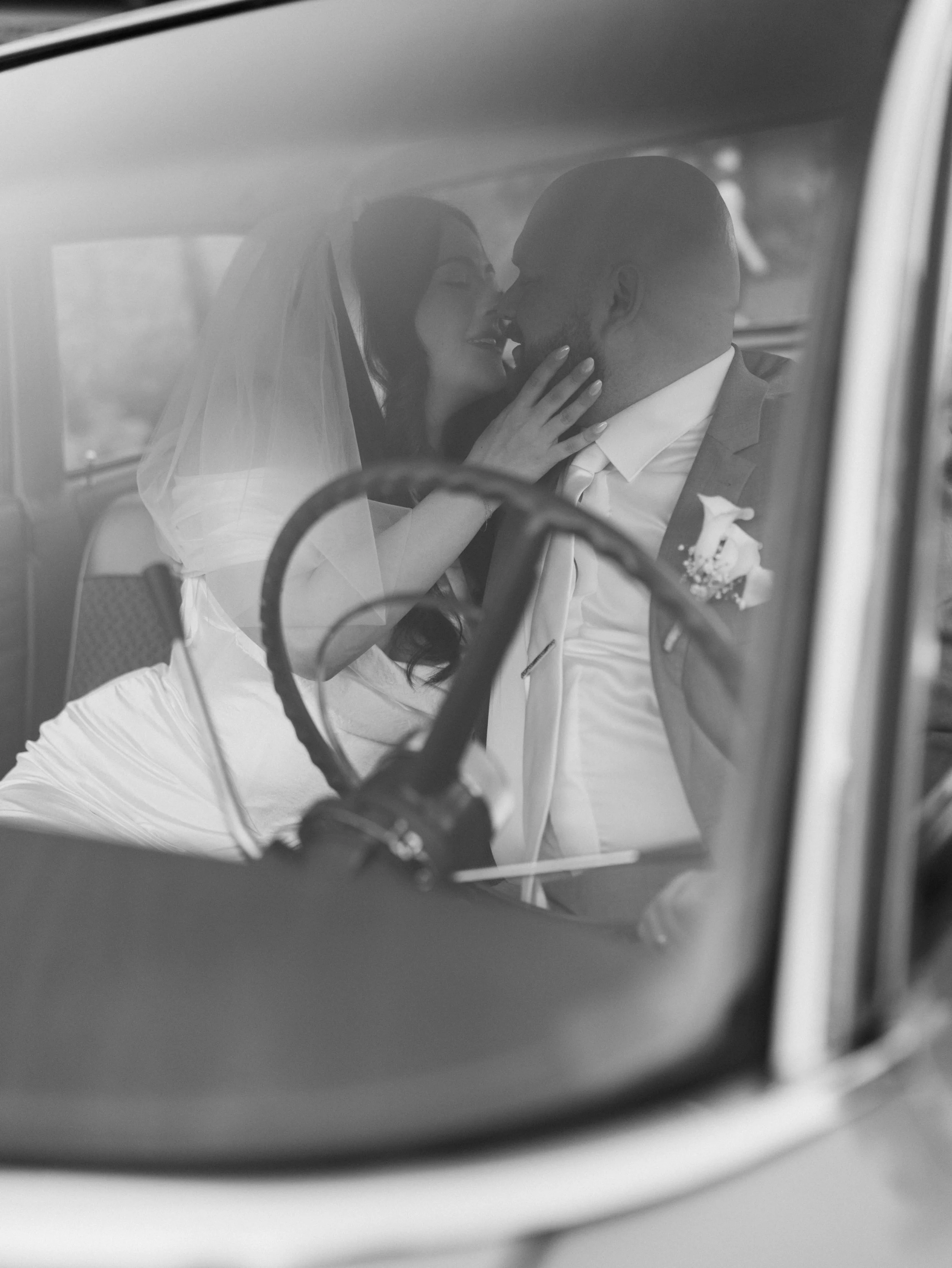 A bride and groom sitting inside a vehicle, sharing a tender moment, with the bride touching the groom's face and both smiling.