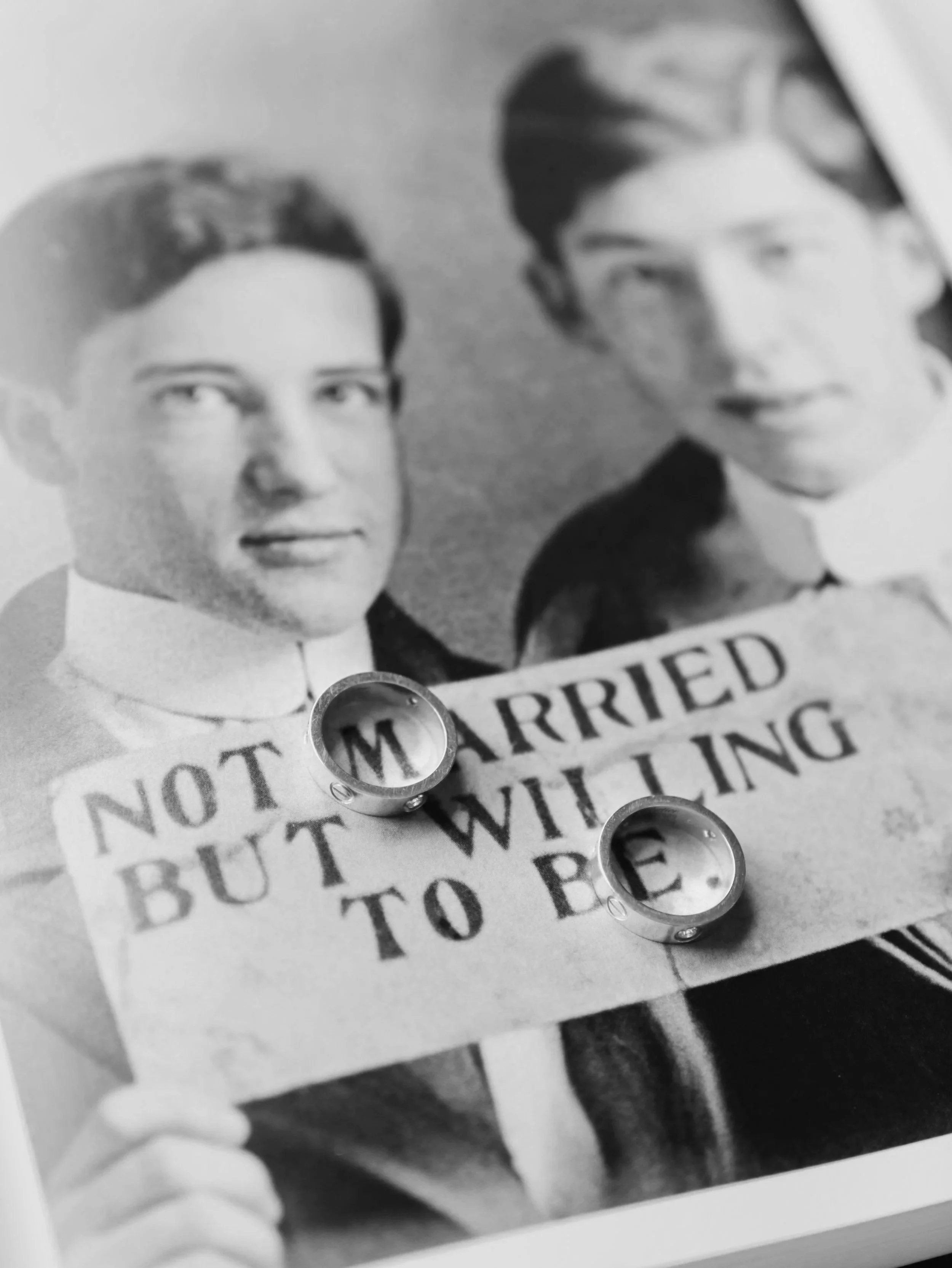 A black and white photo of a couple holding a sign that reads 'Not Married But Willing To Be.' Two wedding rings are placed on top of the sign.